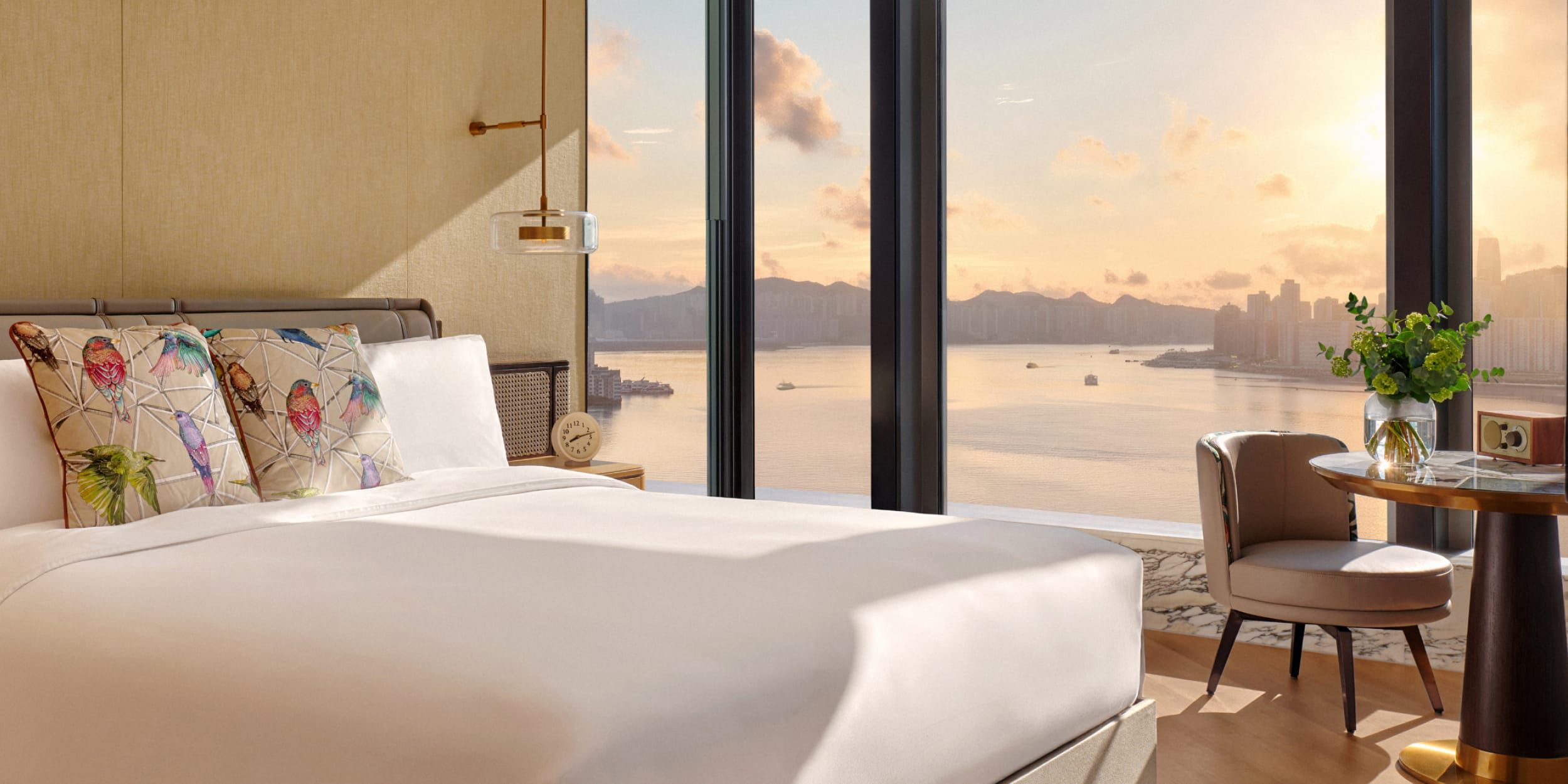 Modern hotel room at Kimpton Tsim Sha Tsui in Hong Kong featuring large windows overlooking a waterfront city at sunset and a neatly made bed