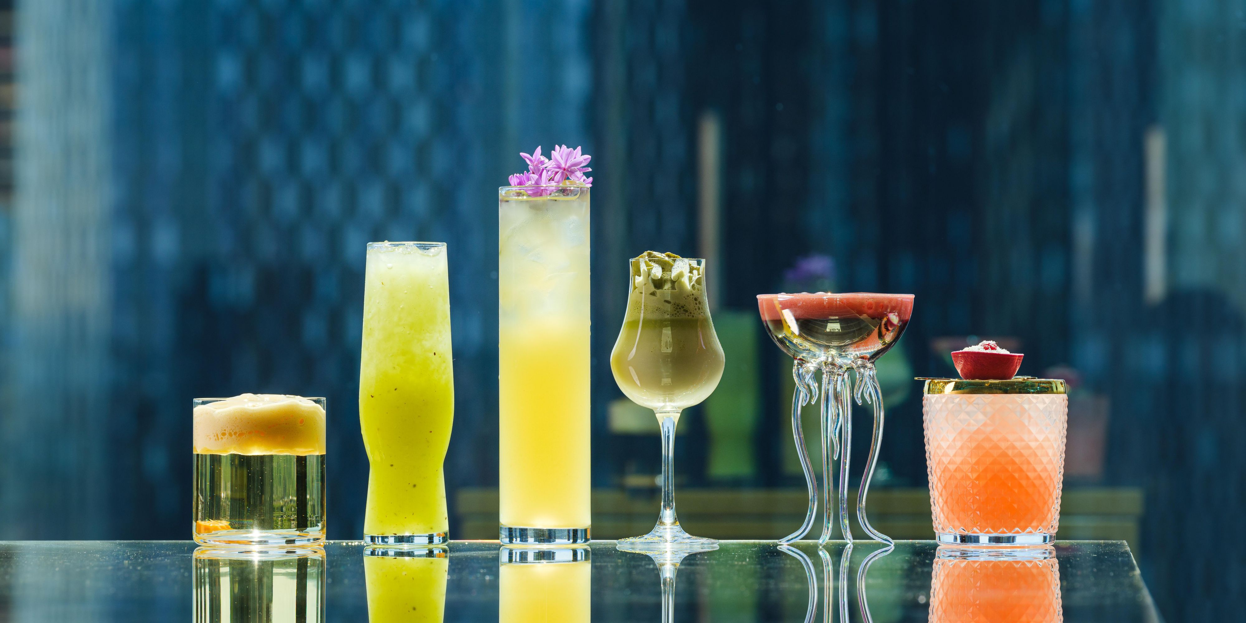 six colourful cocktails lined up across a bar