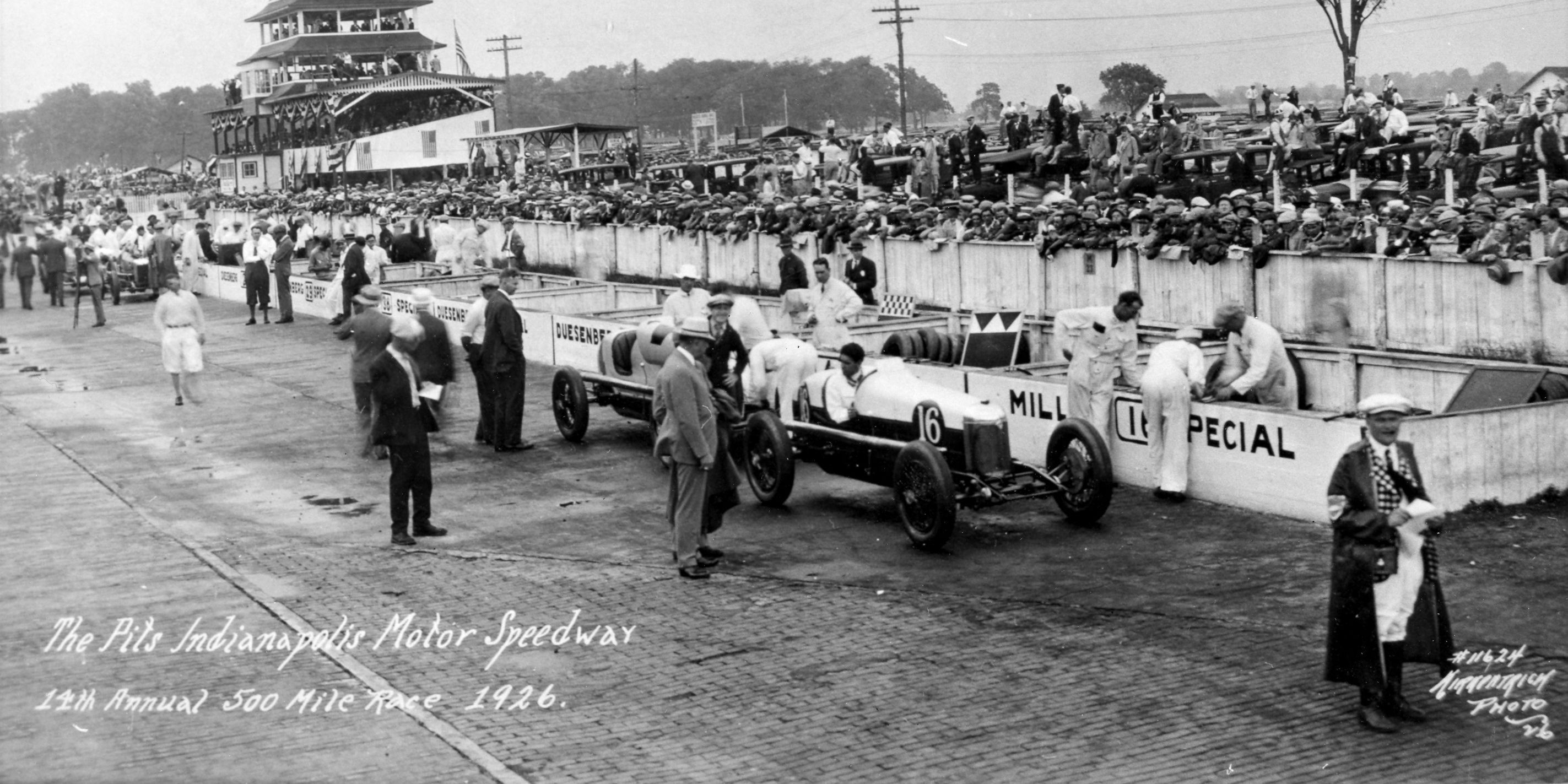 Historic black-and-white photograph of race cars, spectators, and pit crews at the Indianapolis Motor Speedway during the 1926 Indianapolis 500.