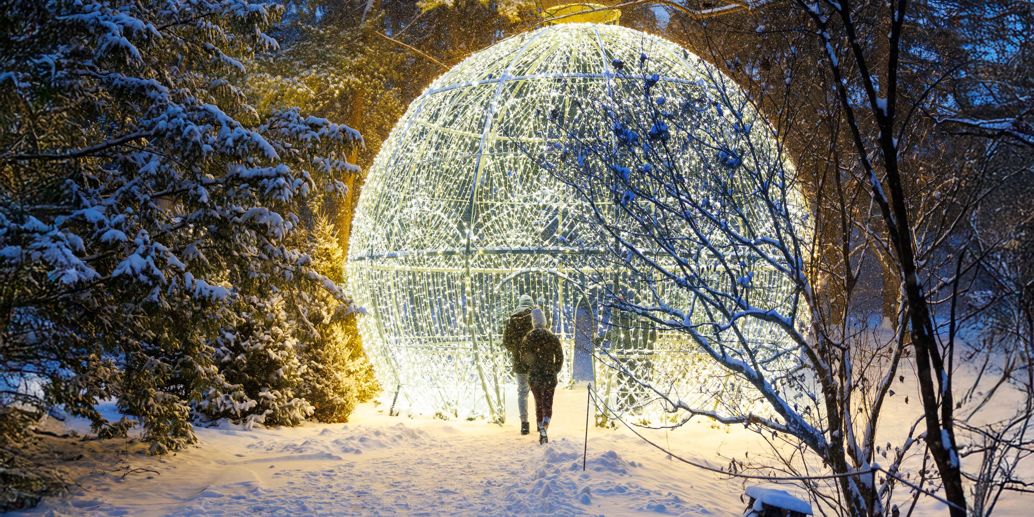 Winterlights display at Newfields featuring a glowing ornament sculpture surrounded by snow-covered trees at dusk.