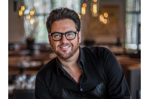 Portrait of a chef standing inside a restaurant dining room with warm ambient lighting.