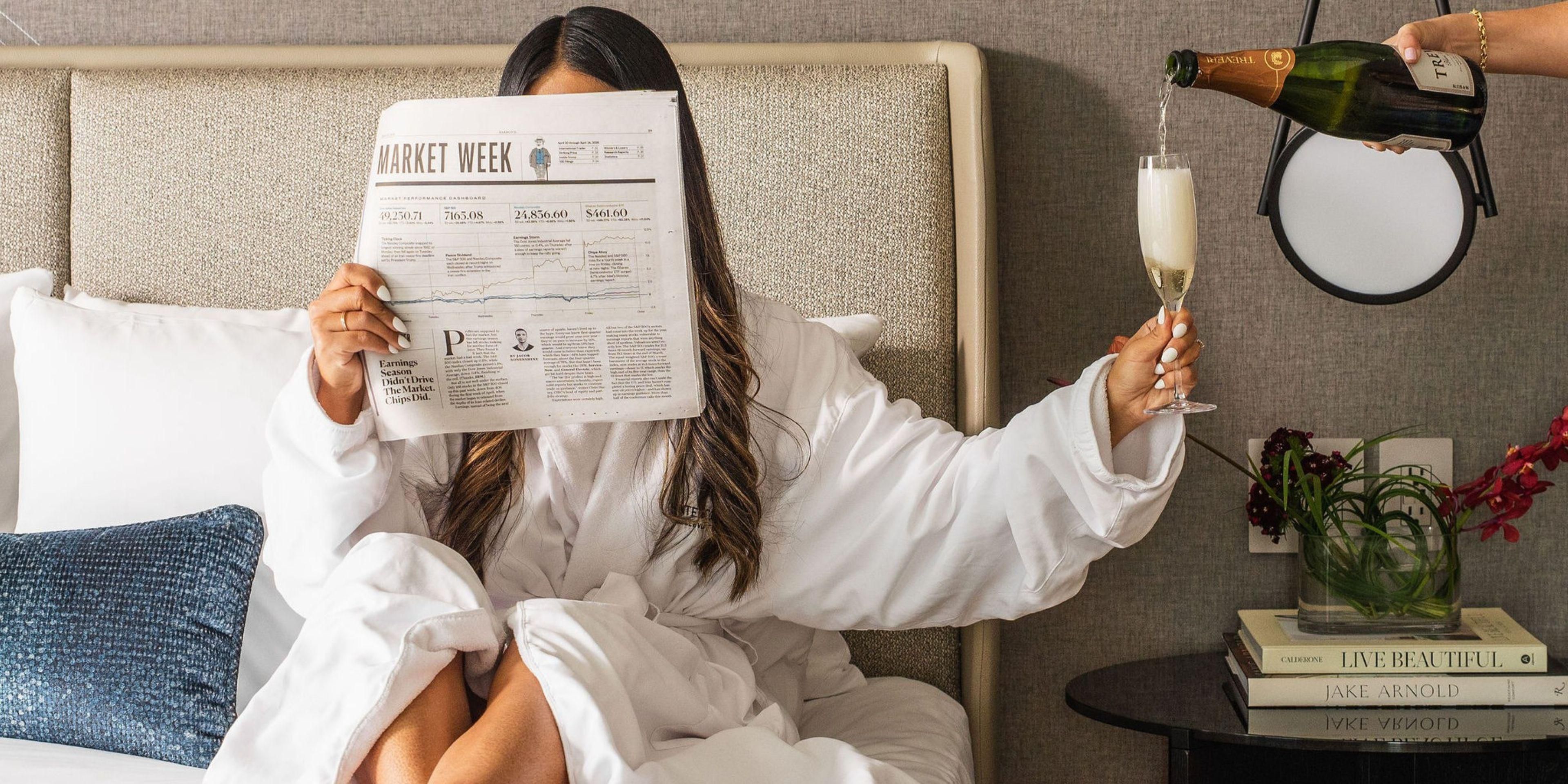 Guest relaxing on hotel bed, reading a newspaper while champagne is being poured for her.