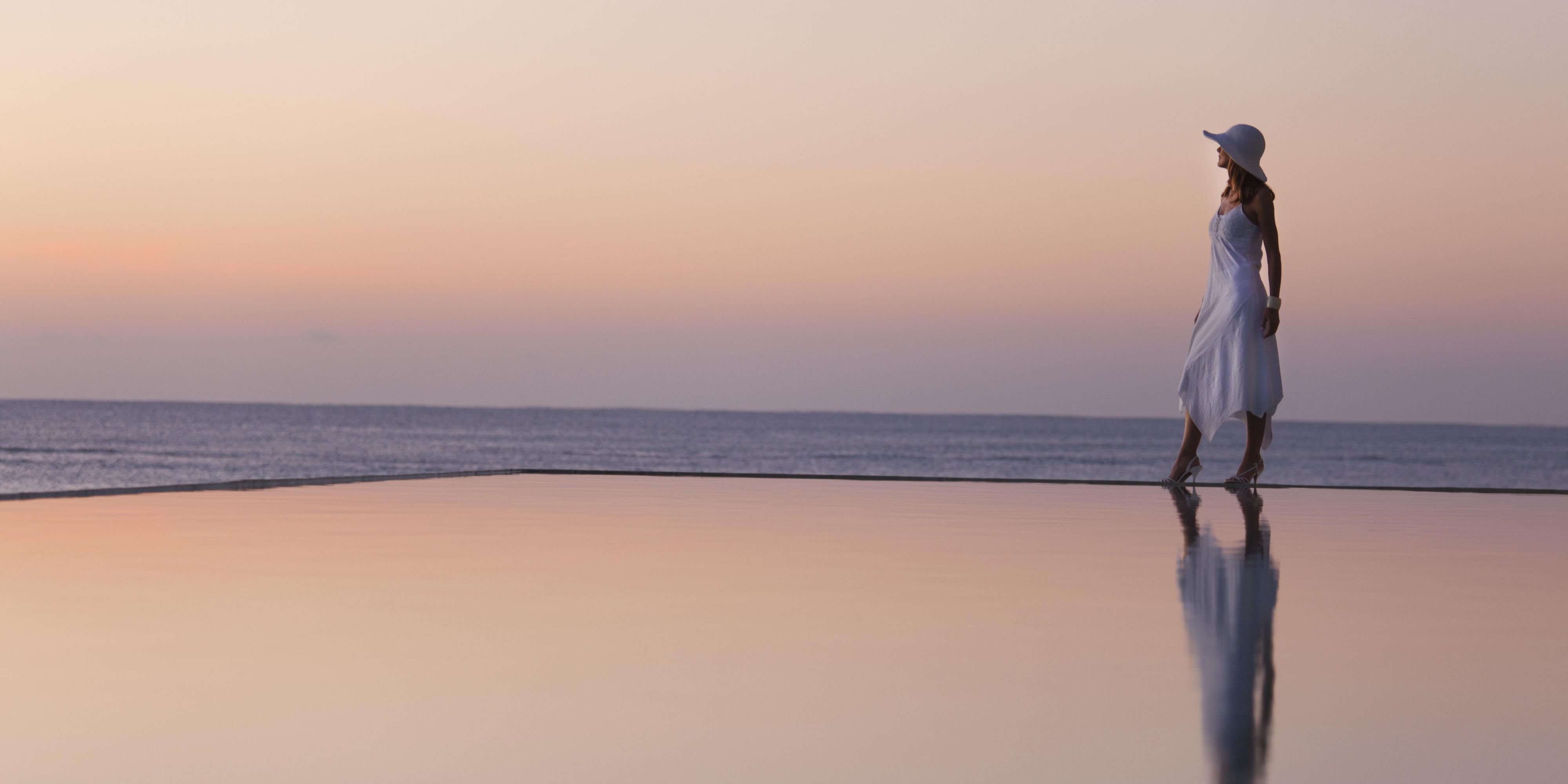 woman dressed in white stood at the edge of the infinity pool in front of the ocean sunset