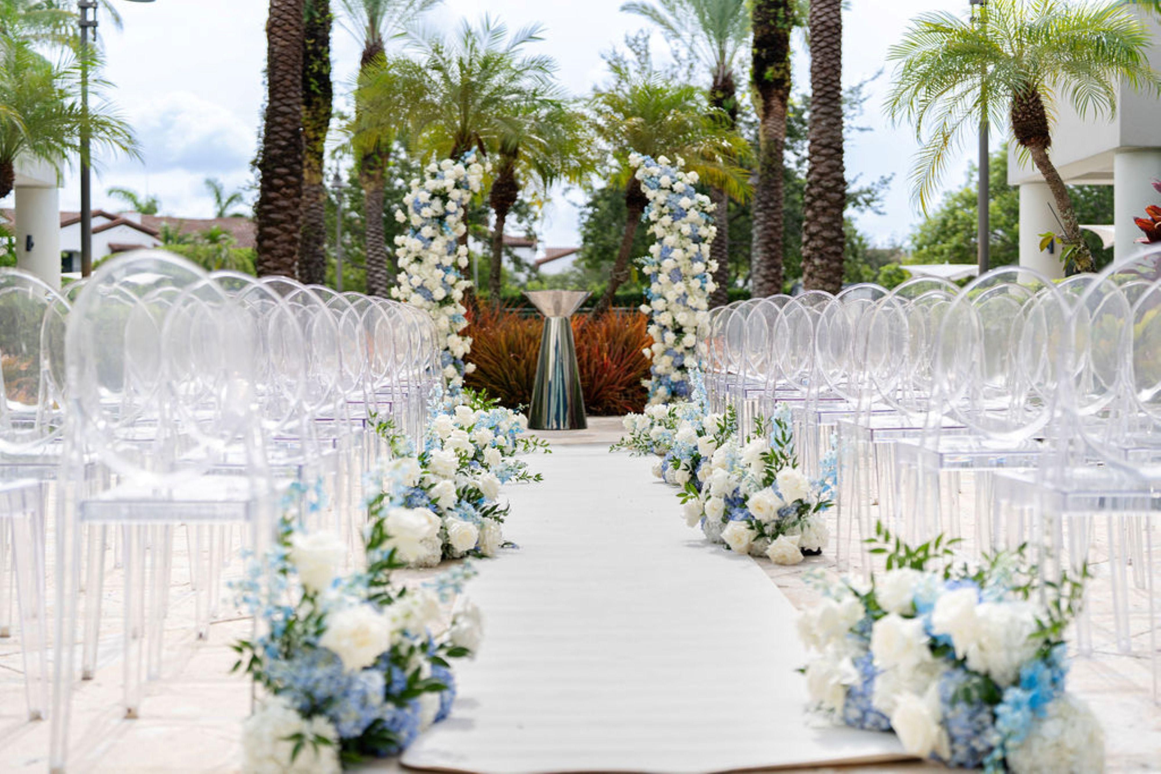 Wedding aisle decorated with white and blue flowers, a floral arch, and clear chairs.