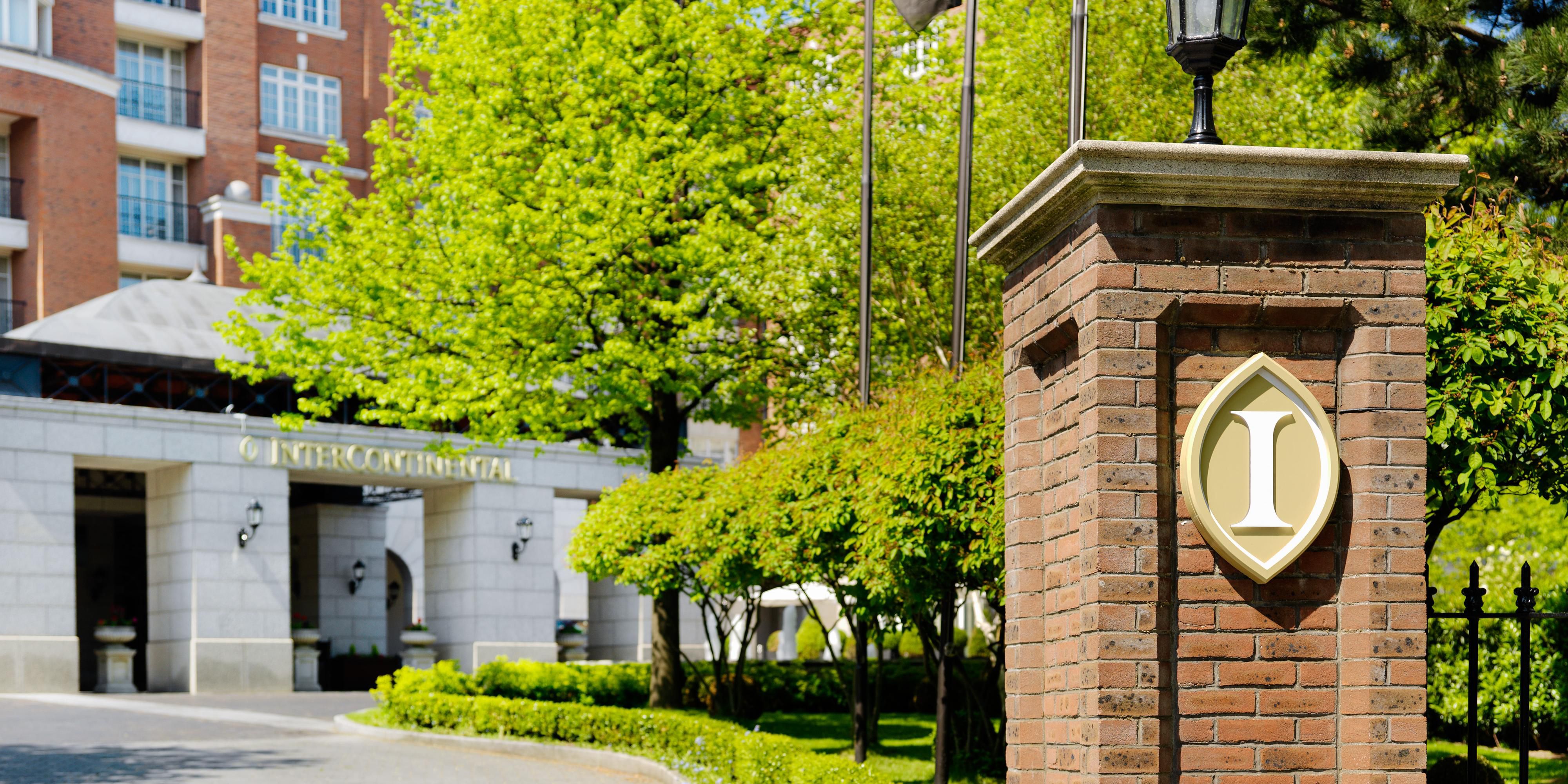 Entrance of a luxury hotel with lush greenery and a brick wall displaying a logo.