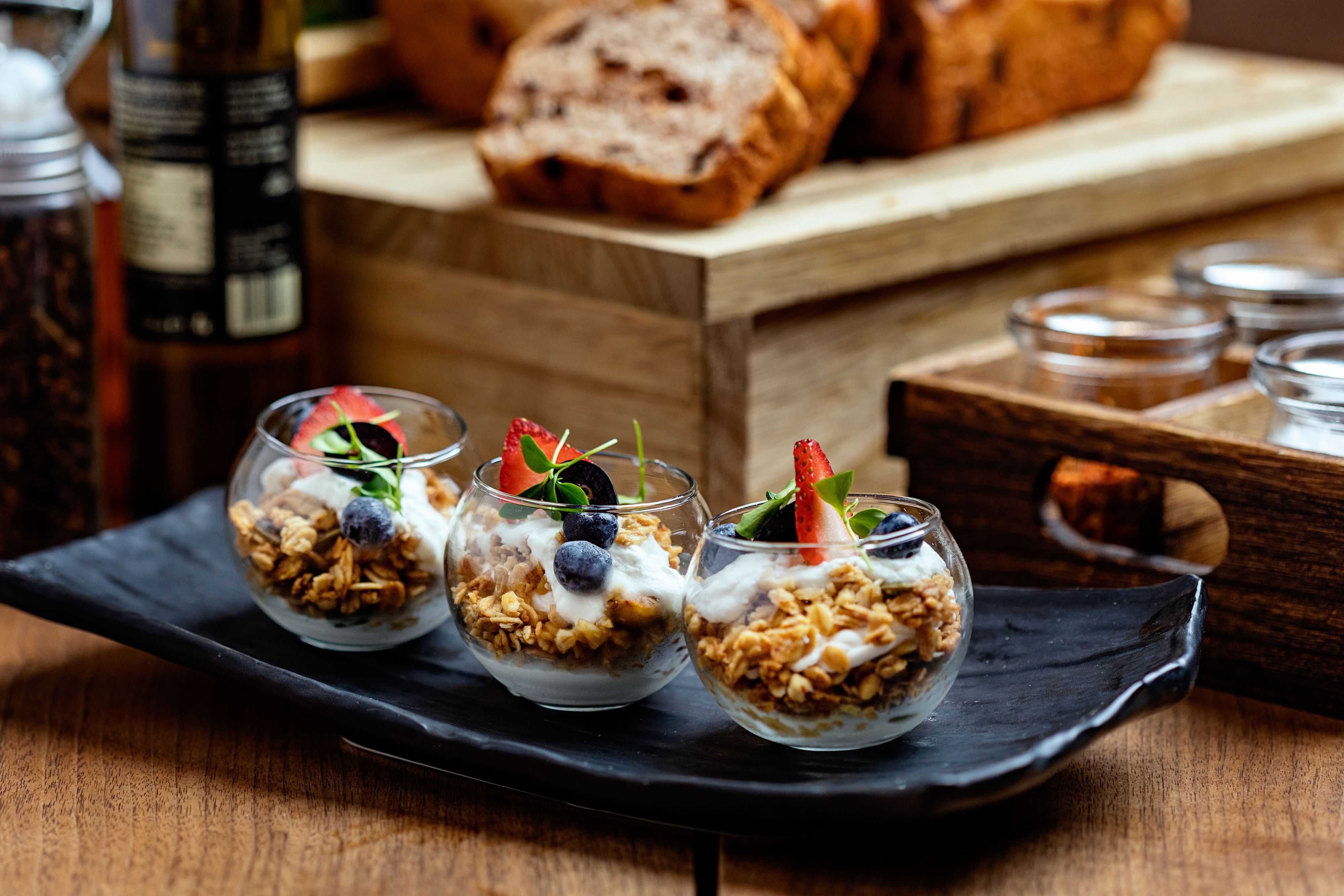 Three small dessert parfaits in glass cups with granola, yogurt, and strawberries on a black tray