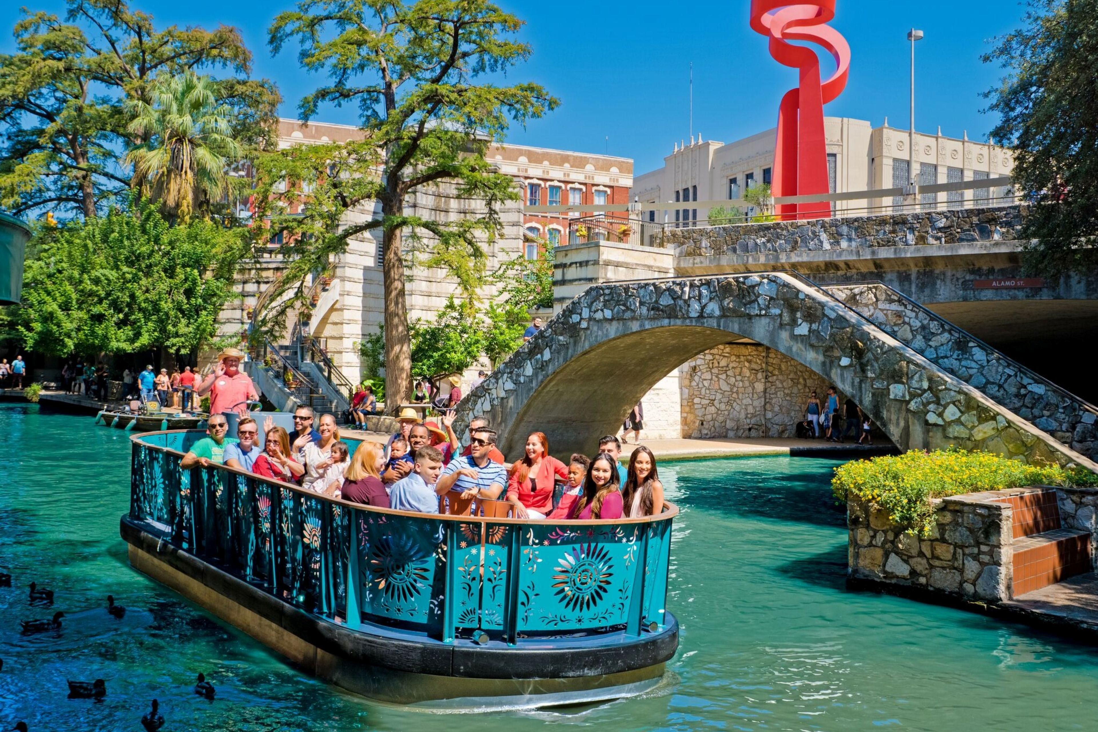 Guests enjoy a scenic river cruise along the San Antonio River Walk, passing beneath a stone bridge and vibrant downtown landmarks.