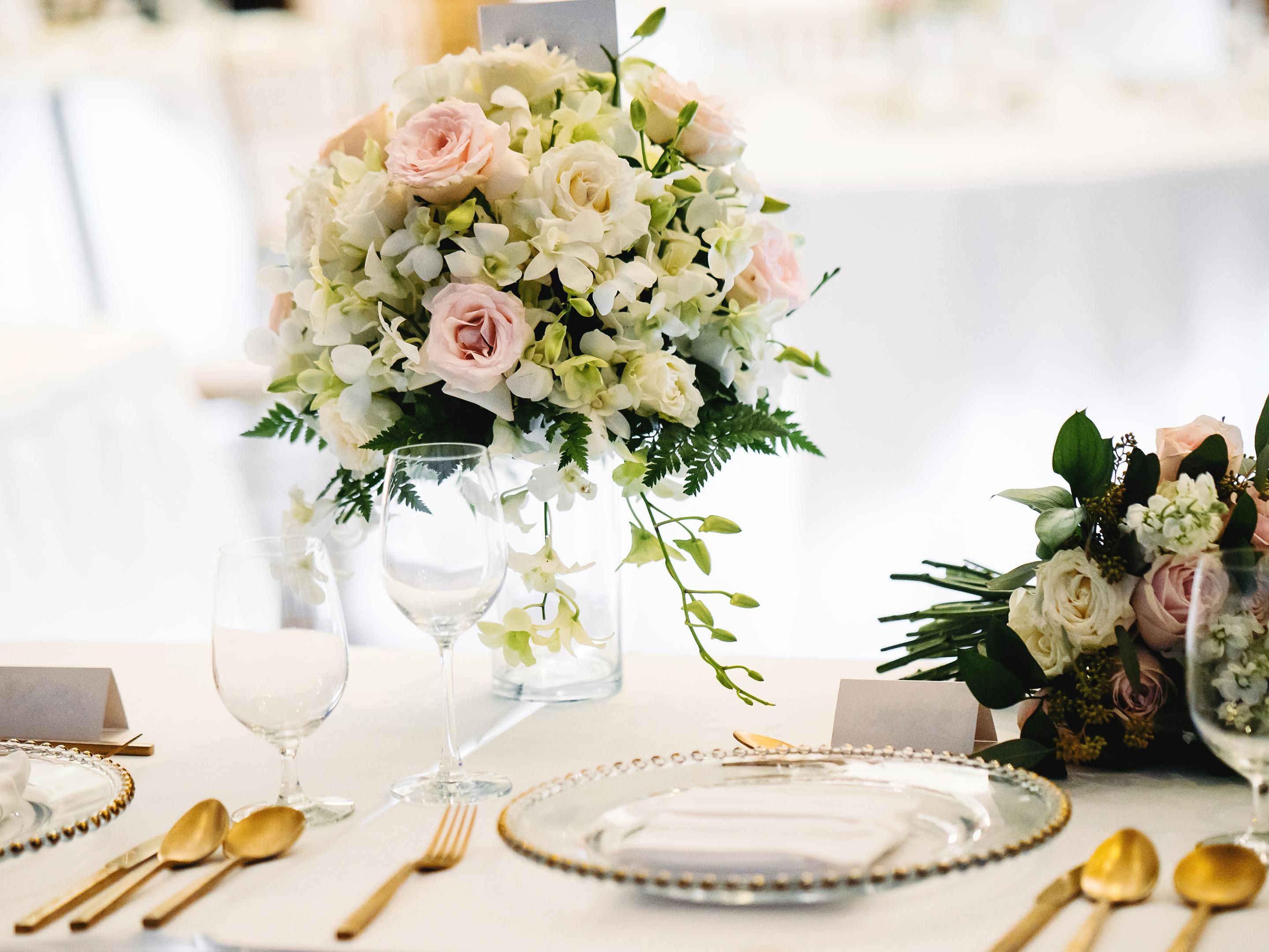 Elegant wedding table setting with white and blush floral centerpieces, gold flatware, and white plates on a cream tablecloth.