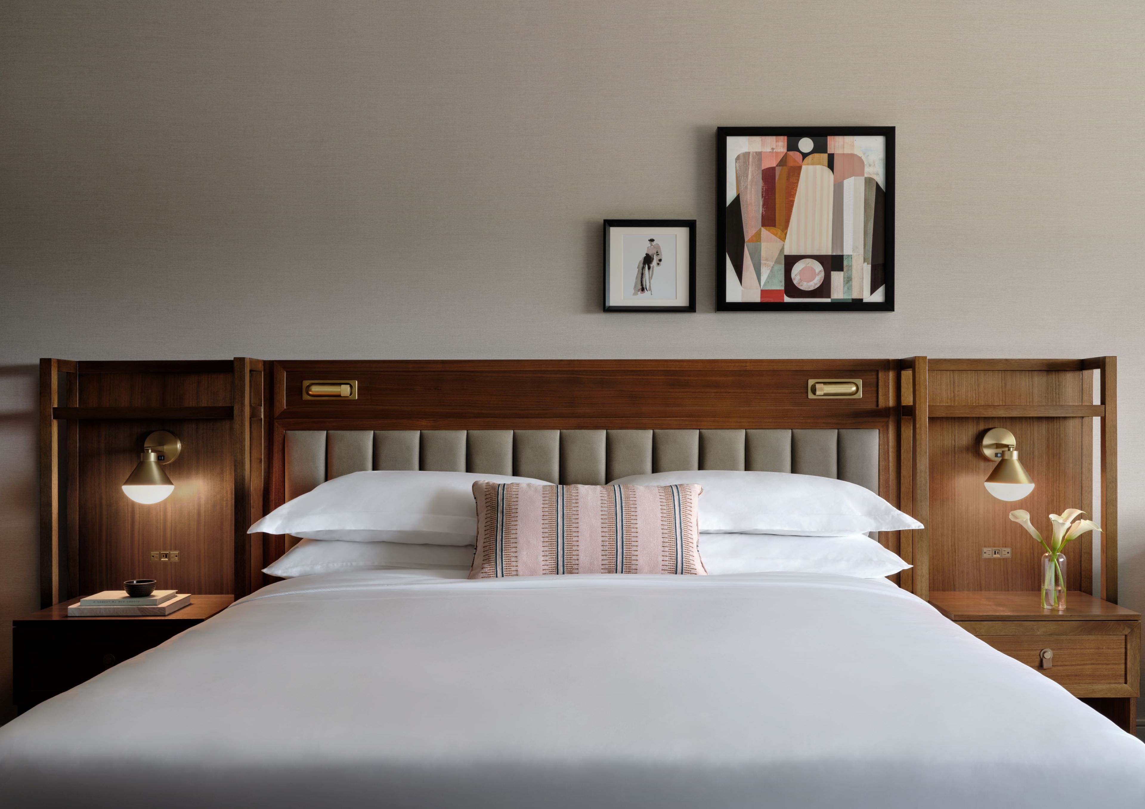 Neatly made bed with white bedding, upholstered headboard, wood nightstands with lamps, and framed artwork above.
