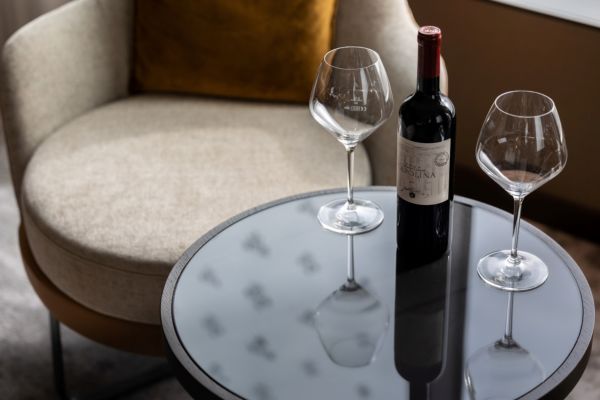Bottle of red wine and two empty wine glasses on a round glass table beside a cushioned chair.