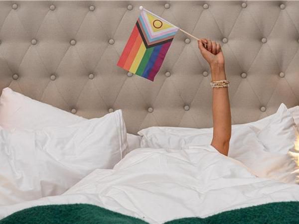A person laying in a double hotel bed holding a pride flag