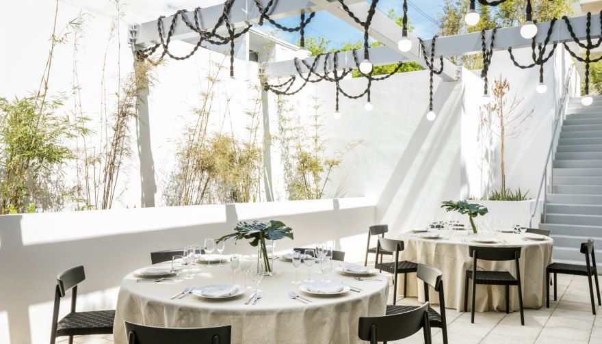 Outdoor event space with bamboo and draped lighting over pergola, white tablecloths, black chairs