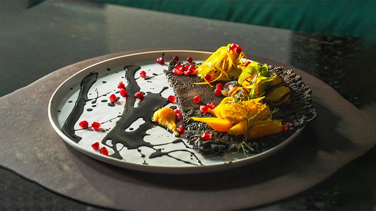 Gourmet dish featuring bright vegetables, pomegranate seeds, and dark sauce on a white plate. 