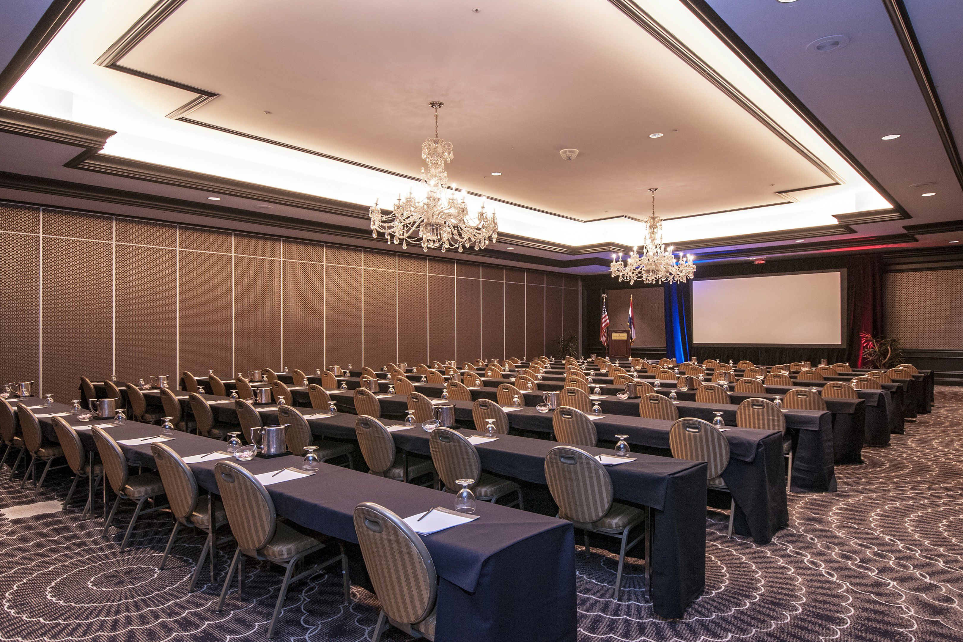Hotel meeting room with classroom-style seating, long tables, notepads, water glasses, and chandeliers overhead.