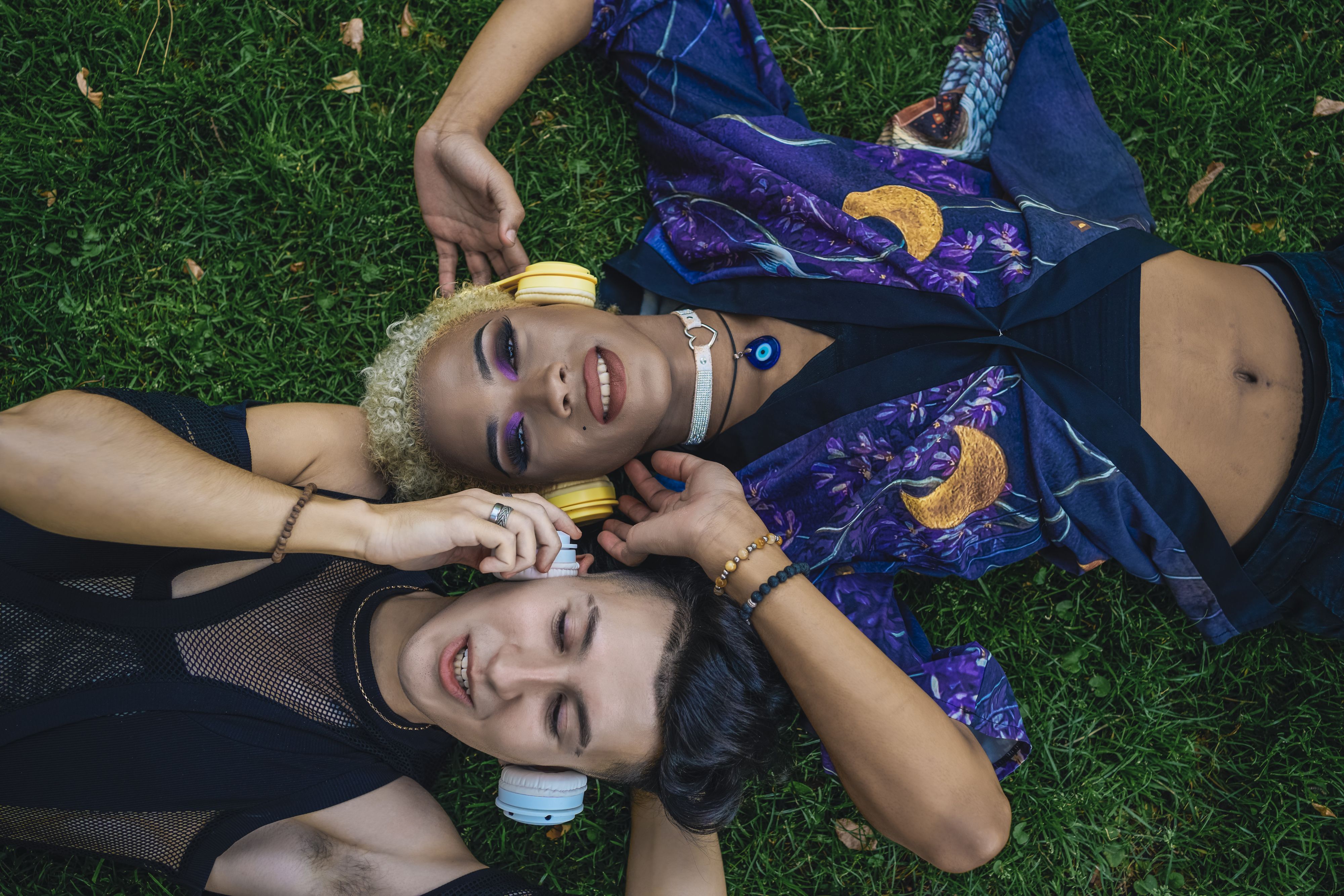 Person lying on grass wearing patterned jacket and holding headphones, with another person beside them, both resting with headphones nearby.