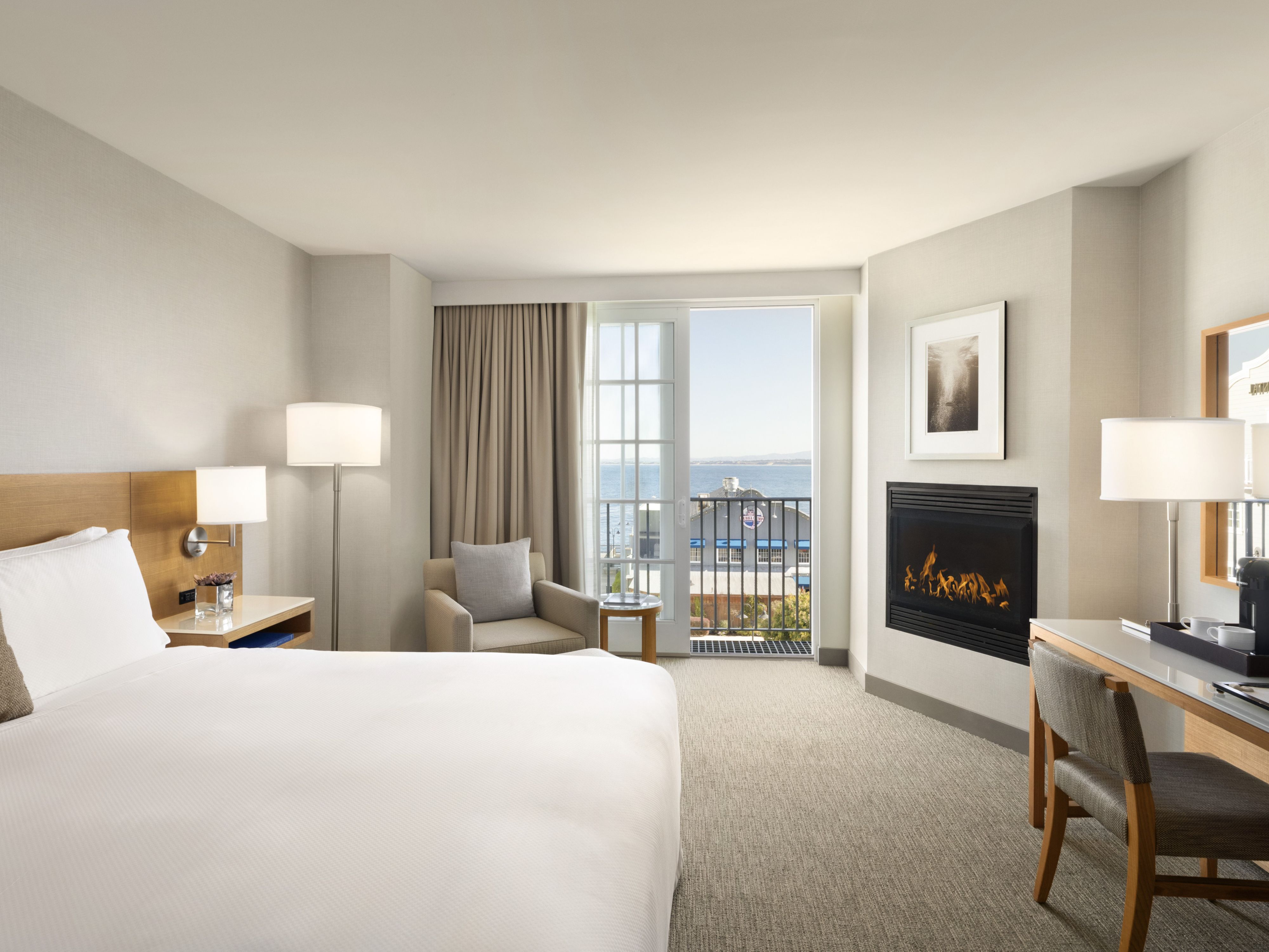 Spacious guest room featuring a king bed, gas fireplace, work desk with coffee maker, upholstered chair, and a private balcony with partial ocean views through glass doors.