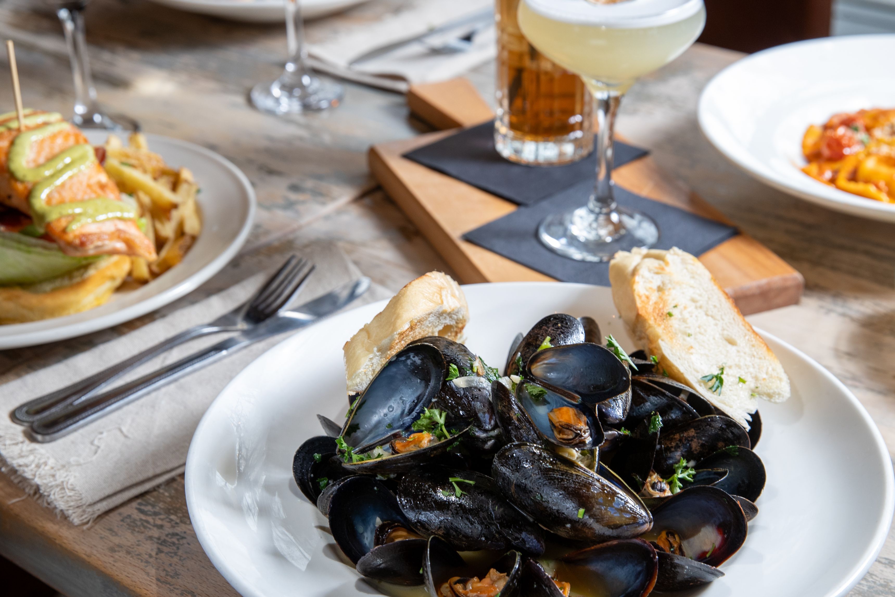 Bowl of steamed mussels with grilled bread and cocktails on a restaurant table.