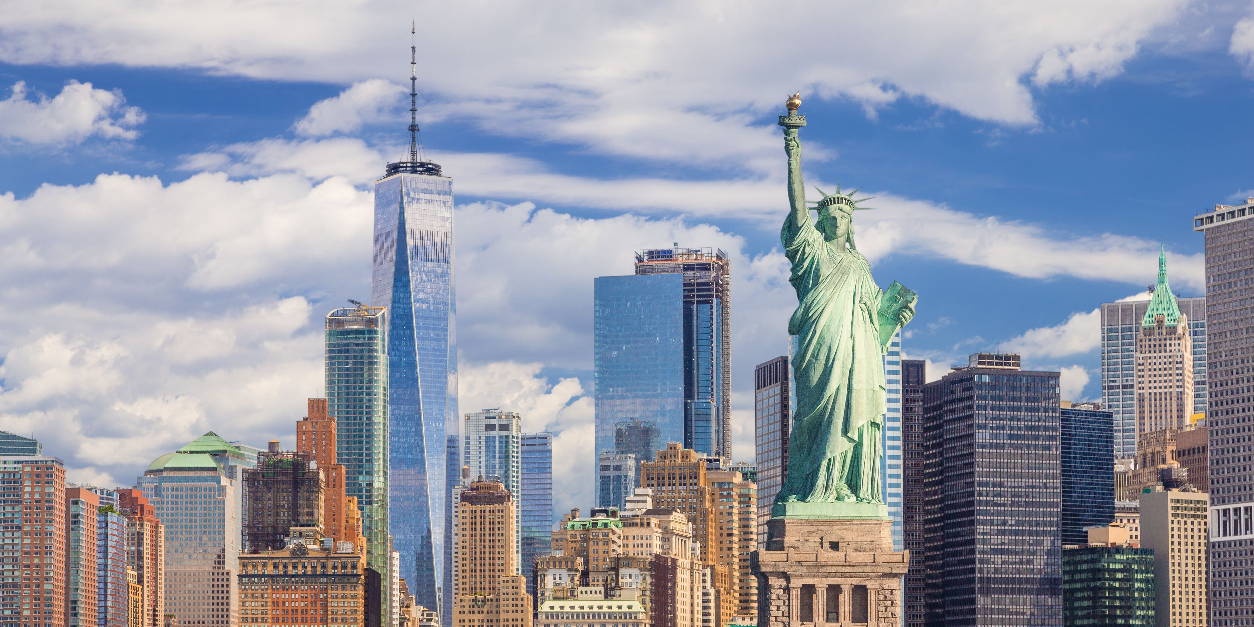 The Statue of Liberty stands before the Lower Manhattan skyline, with One World Trade Center and surrounding skyscrapers rising beneath a bright blue sky filled with billowing white clouds.