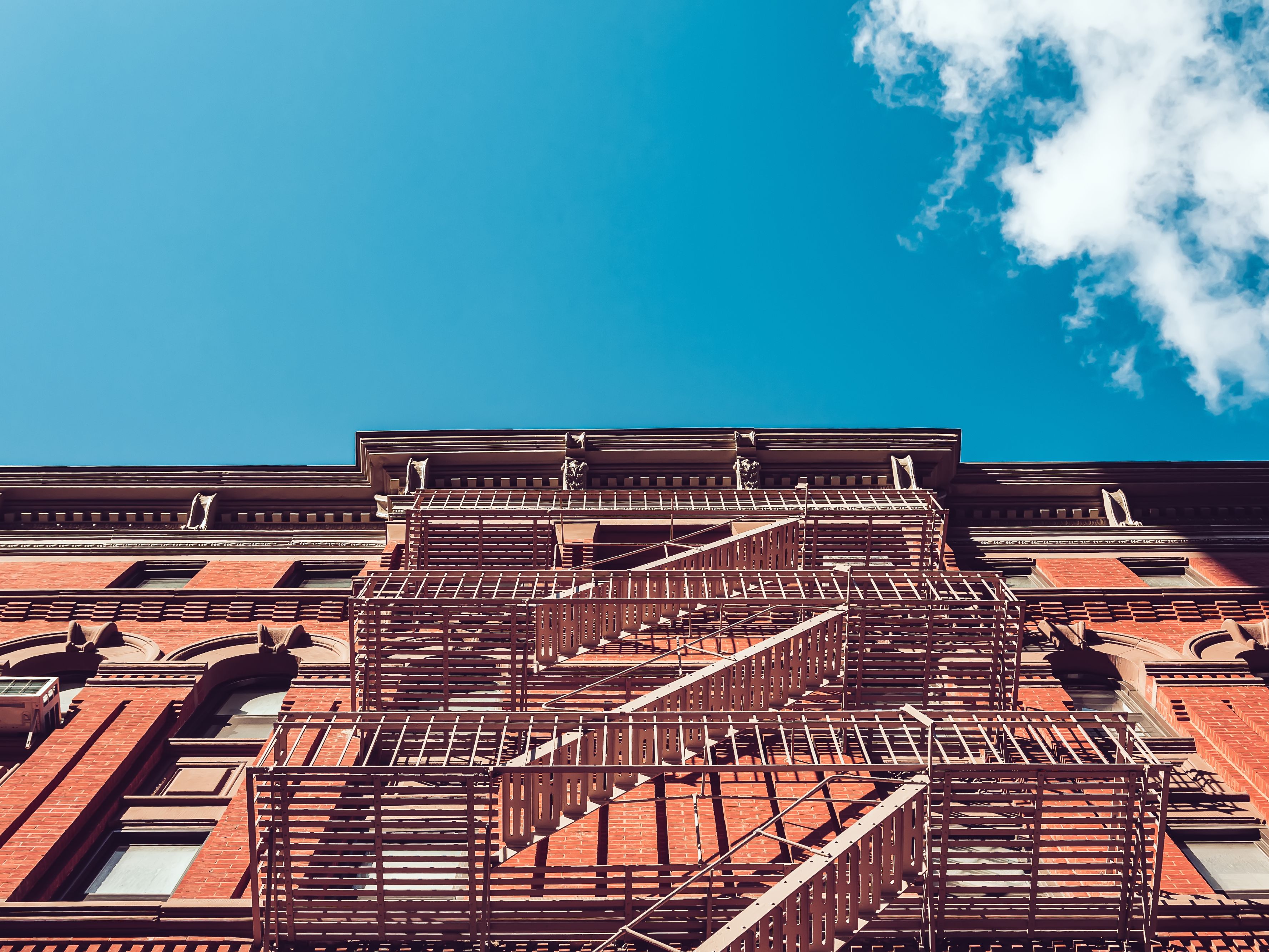 Low-angle view of a historic red brick building with an exterior metal fire escape zigzagging up the façade beneath a bright blue sky with scattered white clouds.