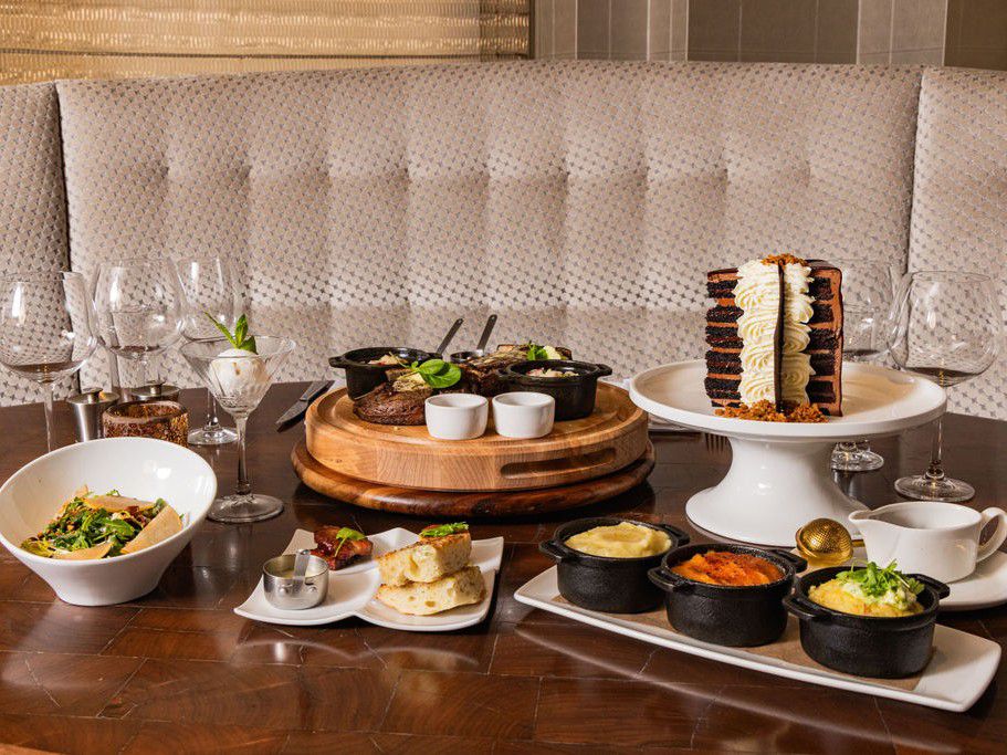 A full restaurant table spread with plated steak, sides, desserts, and drinks arranged in a booth seating area.