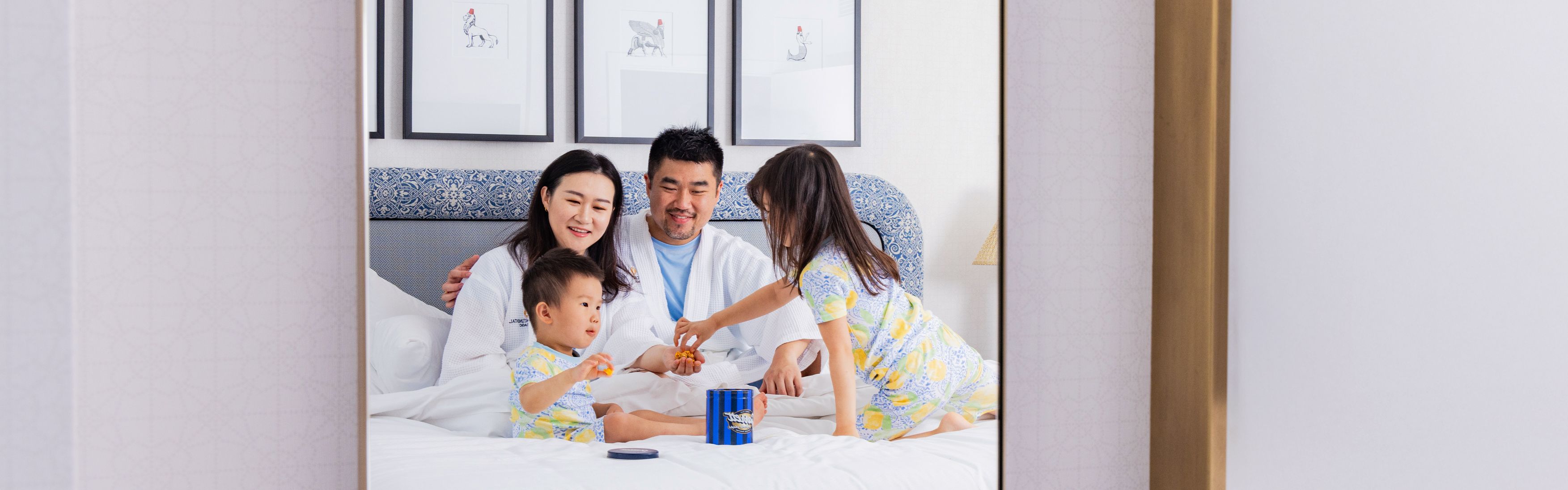 Family gathered on a hotel bed enjoying snacks in a bright guest room.