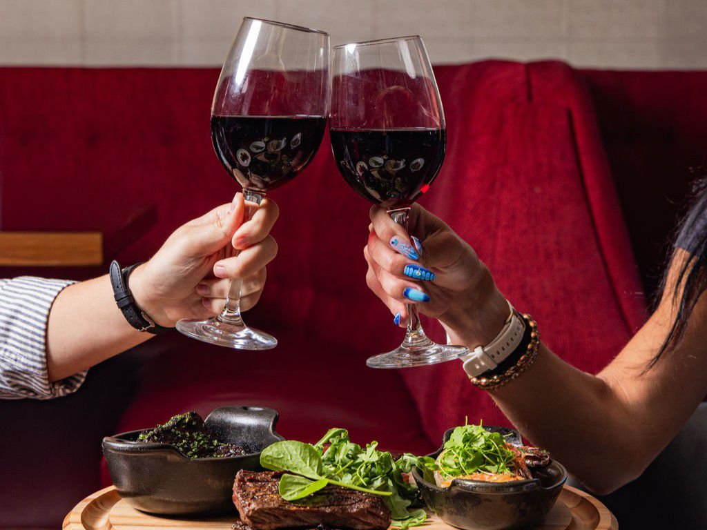 Two hands holding glasses of red wine as they toast over a table with plated steak and side dishes in a restaurant setting.