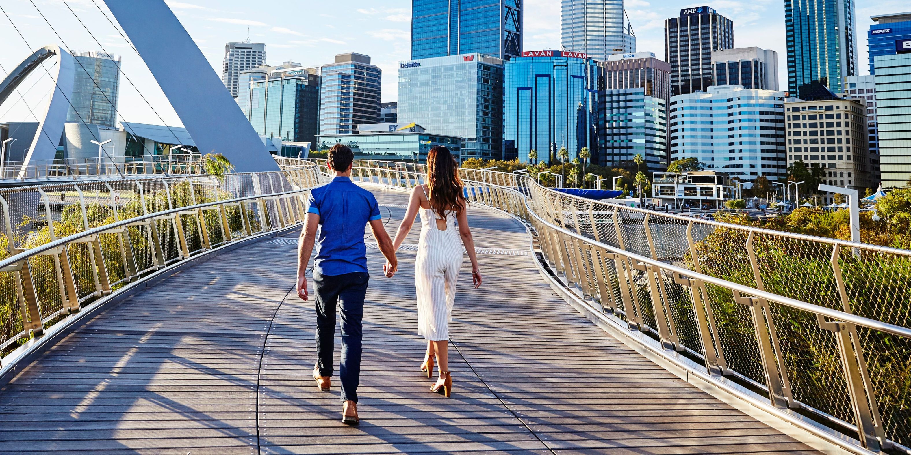 Couple walking across a curved pedestrian bridge with Perth's skyline rising in the background on a clear day.