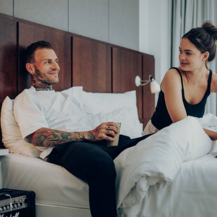 A couple smiling at each other sit on a bed next to an amp