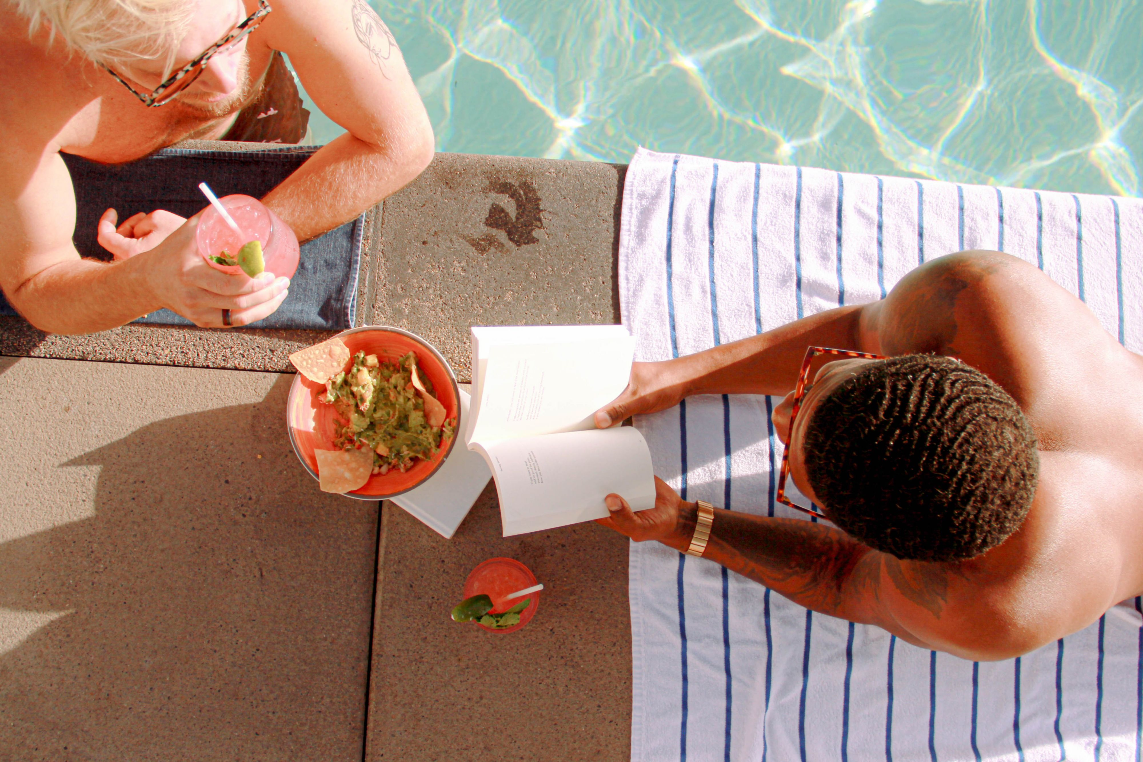 Guests relaxing poolside with cocktails, a book, and light bites beside the swimming pool.