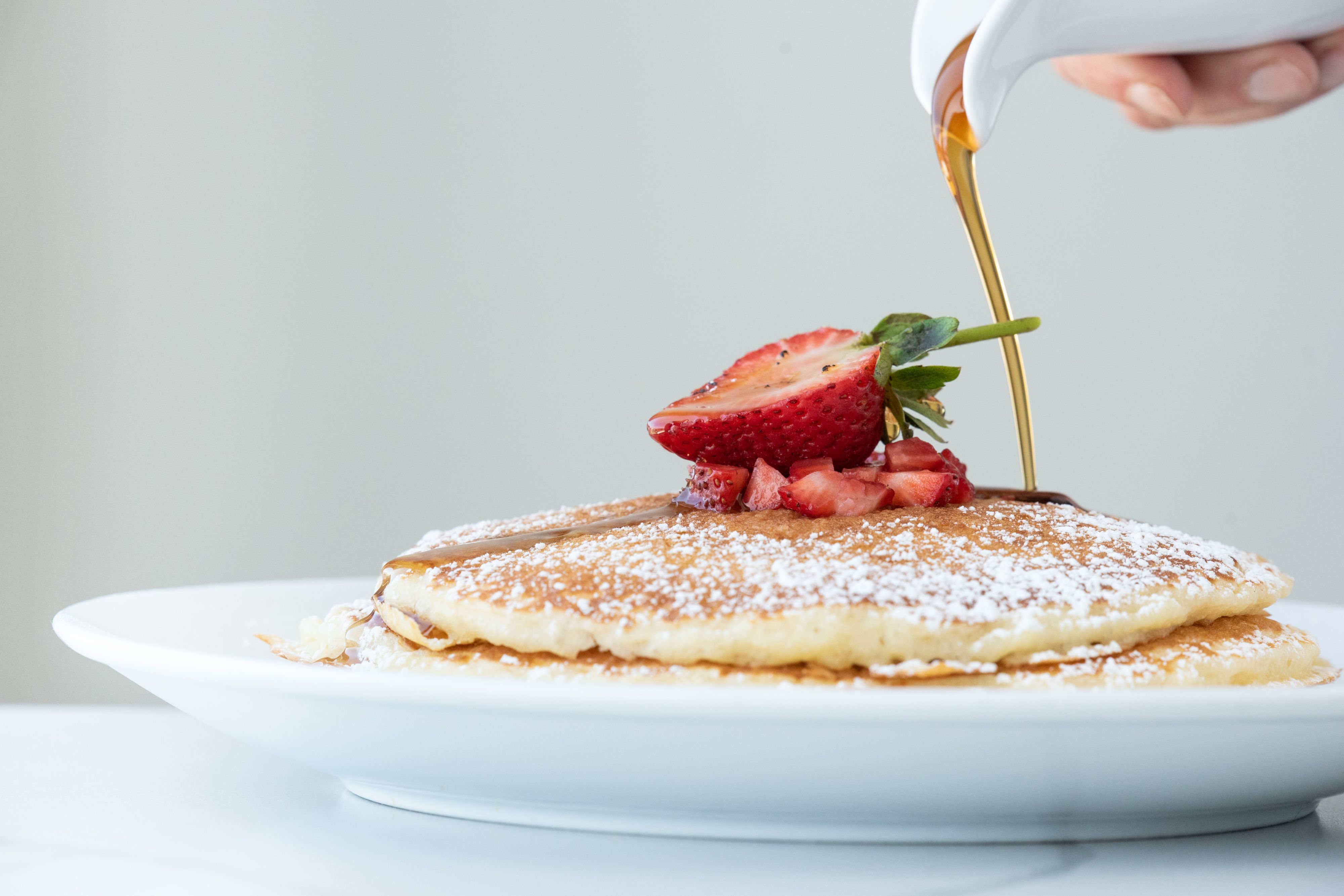 Stack of pancakes topped with fresh strawberries and powdered sugar as syrup is poured over them.