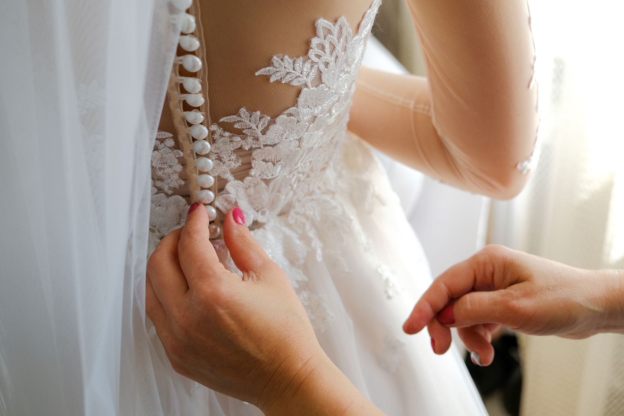 Close-up of hands fastening pearl buttons on a lace wedding dress with sheer detailing.