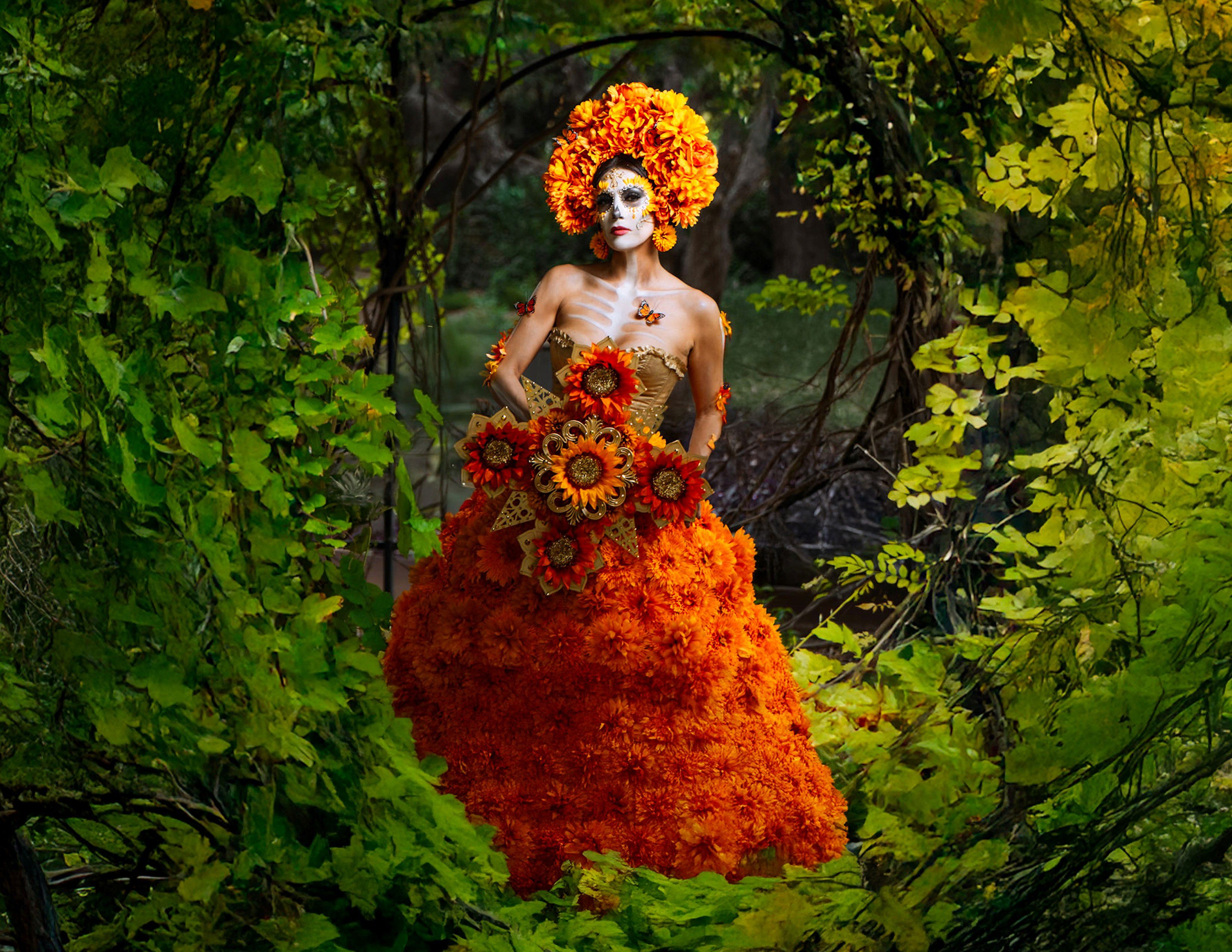 Person in elaborate marigold dress and skull makeup standing in a garden.