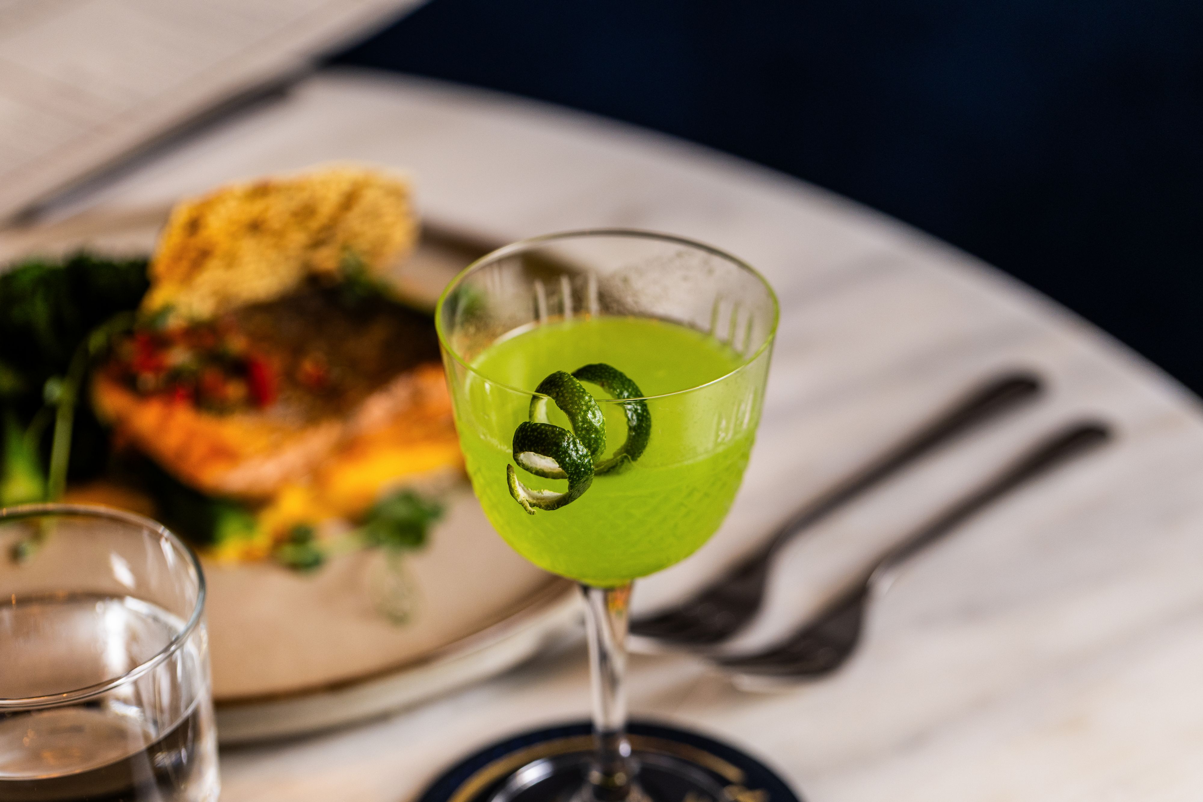 Bright green cocktail garnished with cucumber slices on a marble table beside a plated meal.