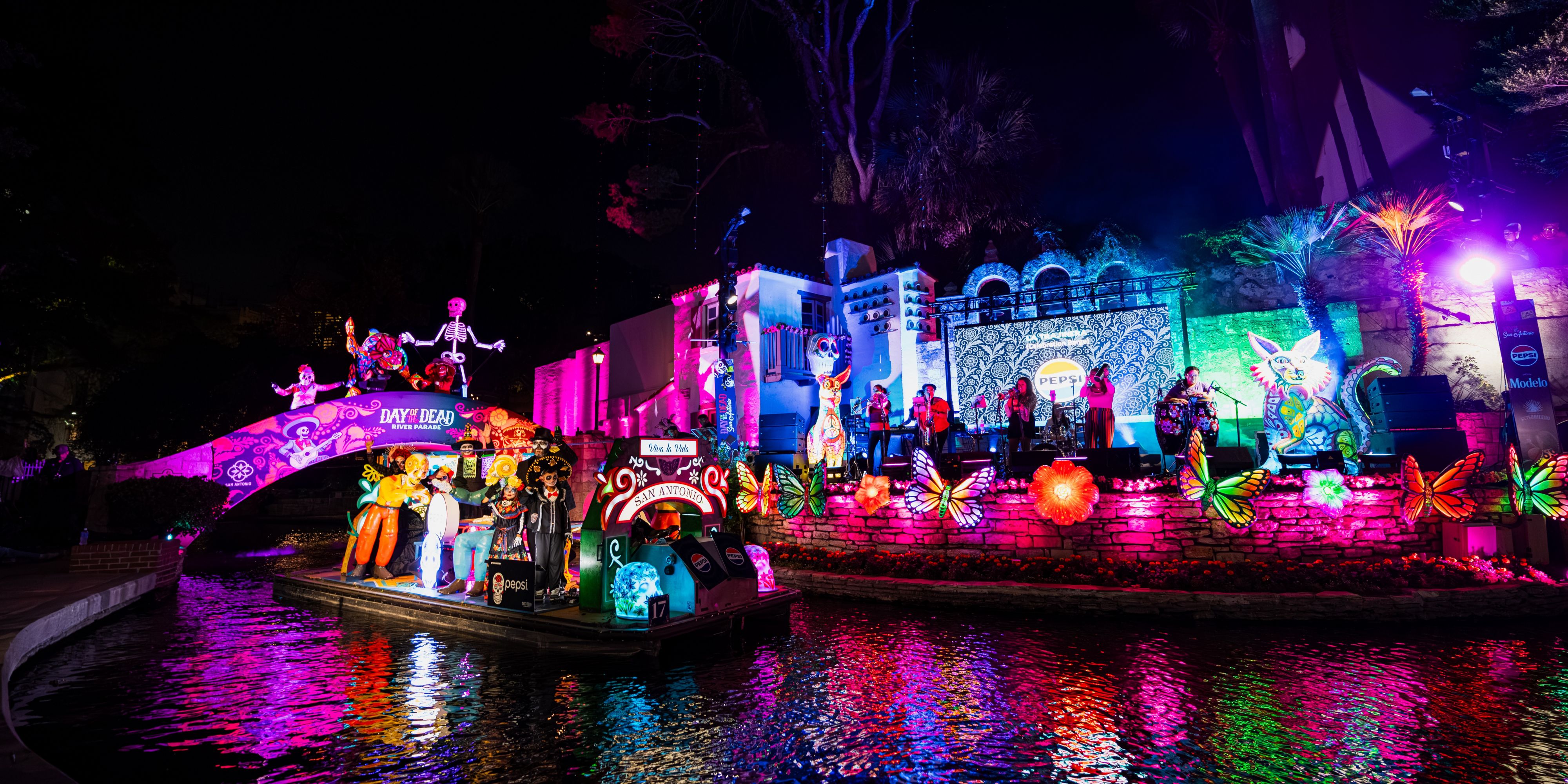 Colorful Día de los Muertos river parade float passing illuminated decorations and live music along the San Antonio River Walk at night.