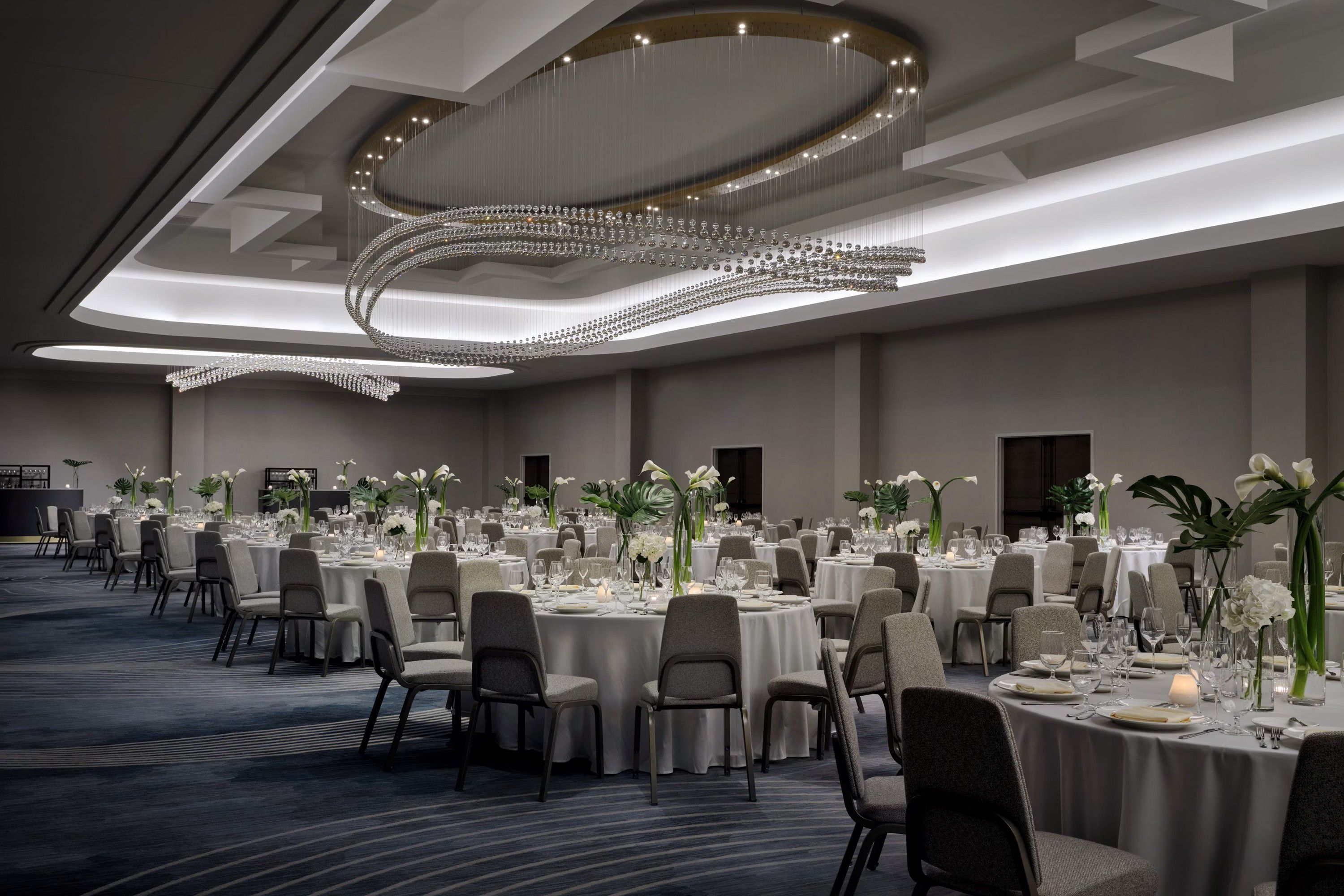Large ballroom set for an event with round tables, floral centerpieces, and modern chandeliers.