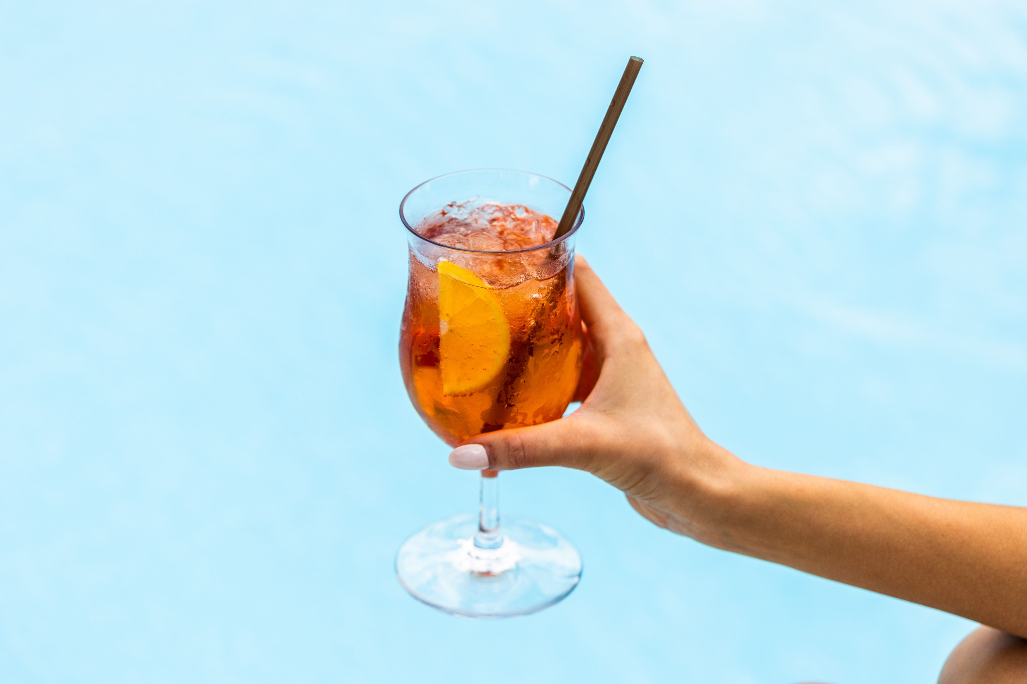 A hand holding an orange cocktail with ice and an orange slice against a blue background.