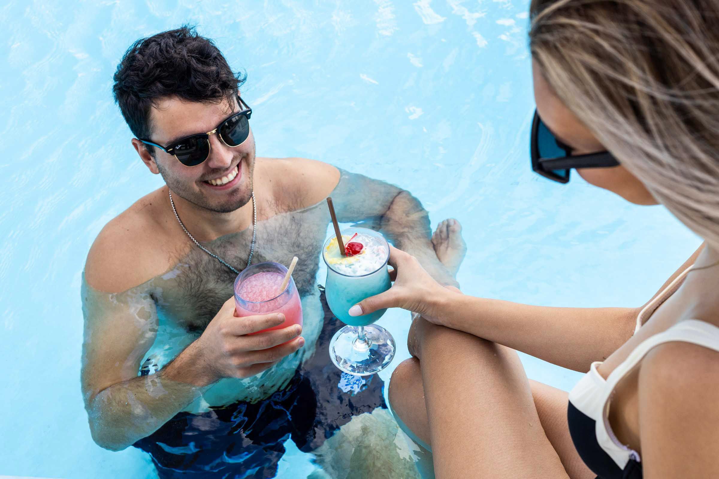Two guests holding colorful cocktails while relaxing in a swimming pool.