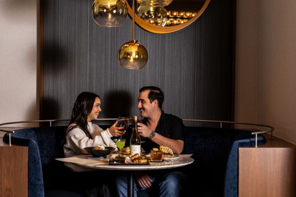 Two people clinking wine glasses at a restaurant table with shared plates and pendant lighting overhead.