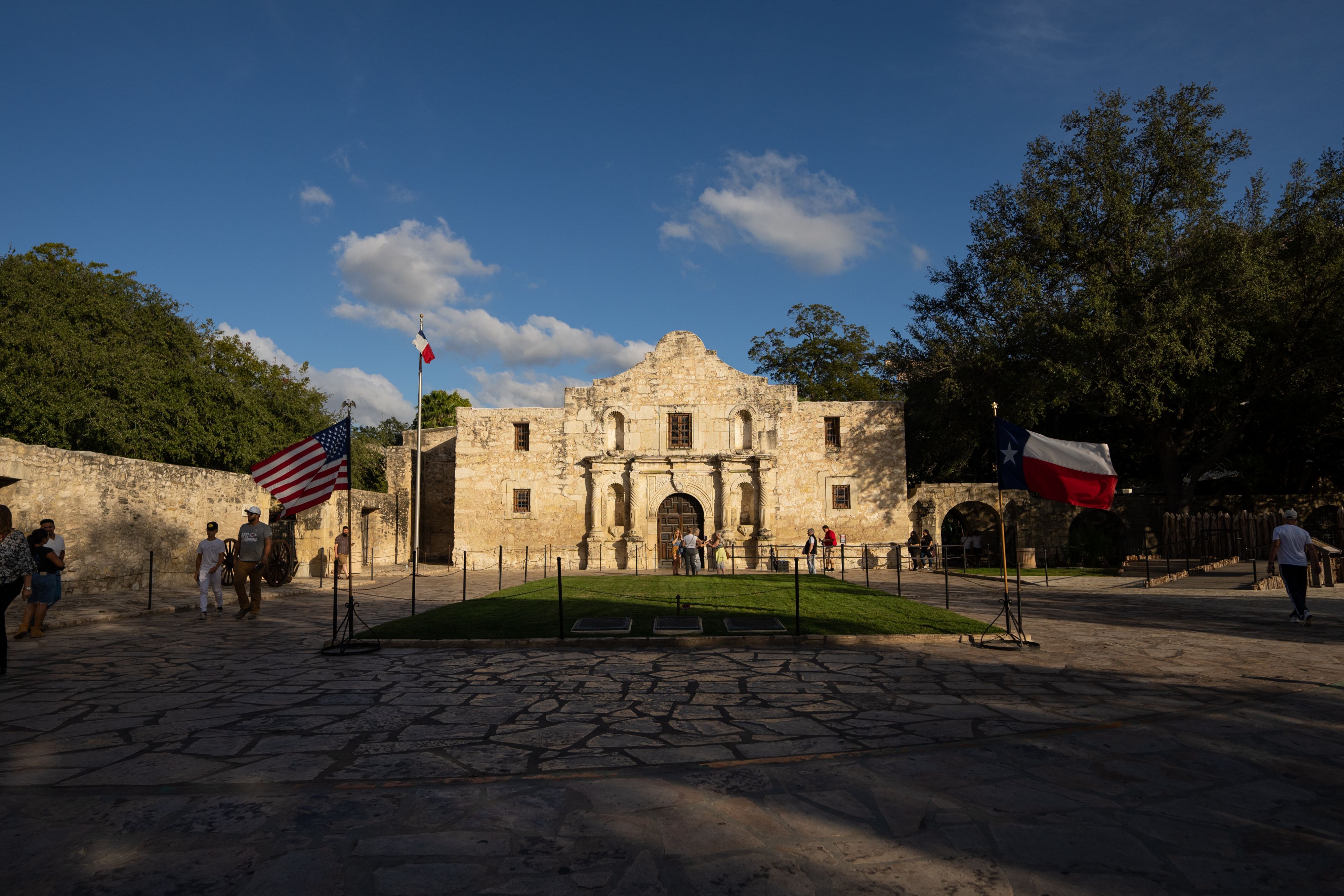 The historic stone façade of the Alamo stands centered in a courtyard, with visitors walking nearby and U.S. and Texas flags displayed on either side under a clear blue sky.