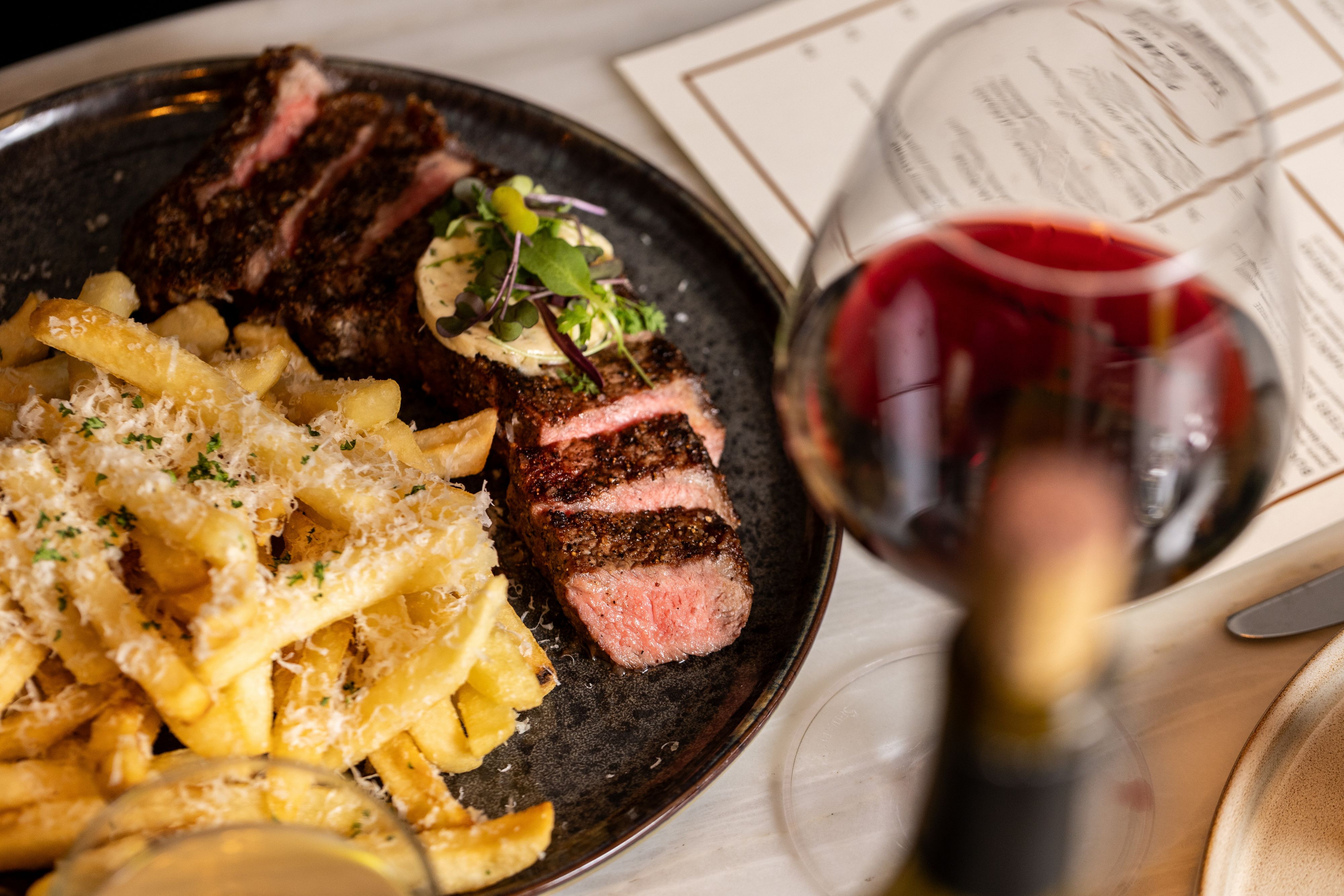 Sliced steak served with parmesan fries on a black plate beside a glass of red wine.