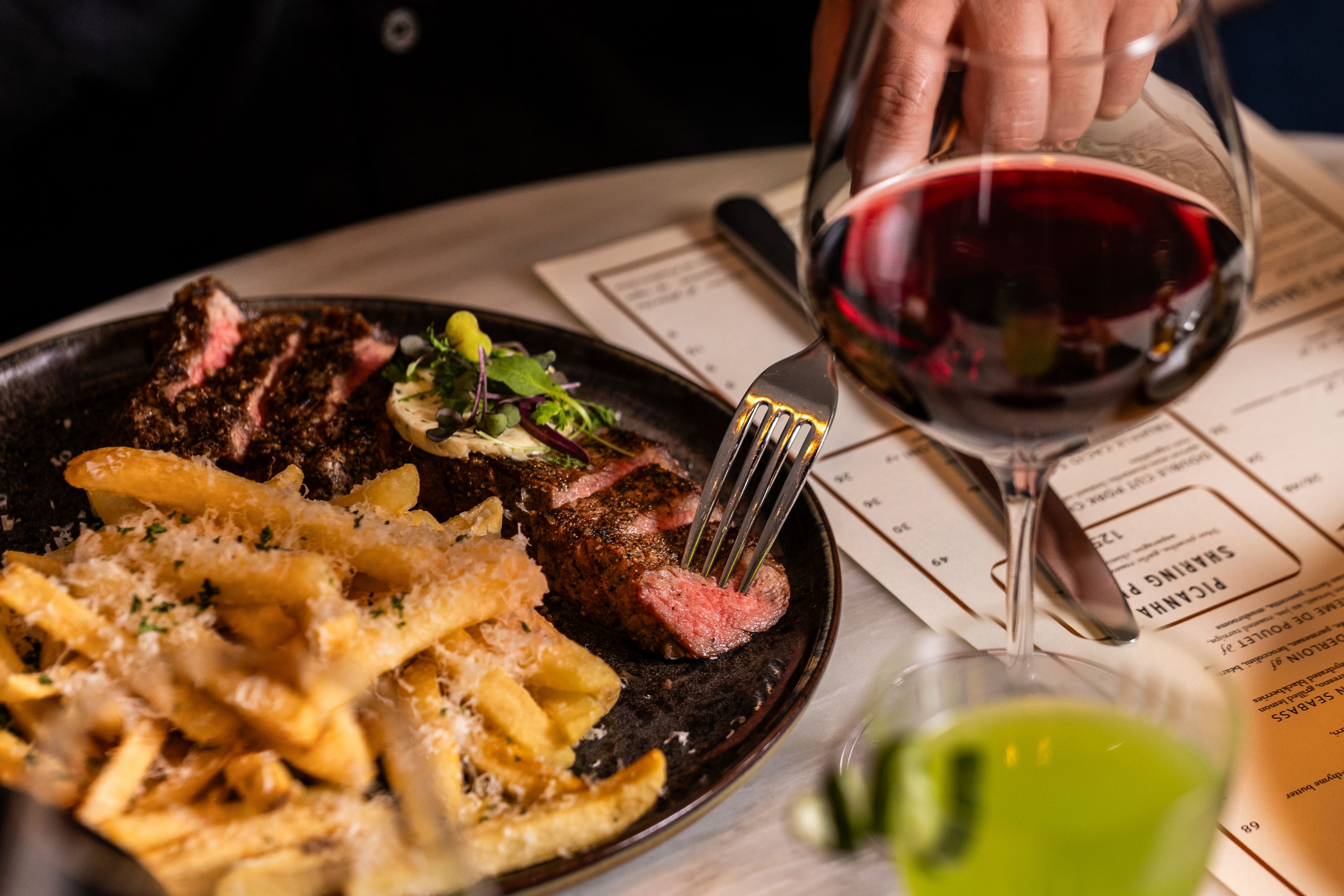 Steak and pasta dish on a table with a glass of red wine being served.