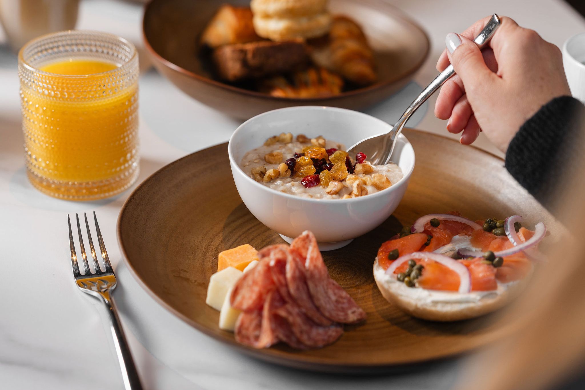 Plate with a salmon bagel, oatmeal bowl, and meat and cheeses next to a glass of orange juice.
