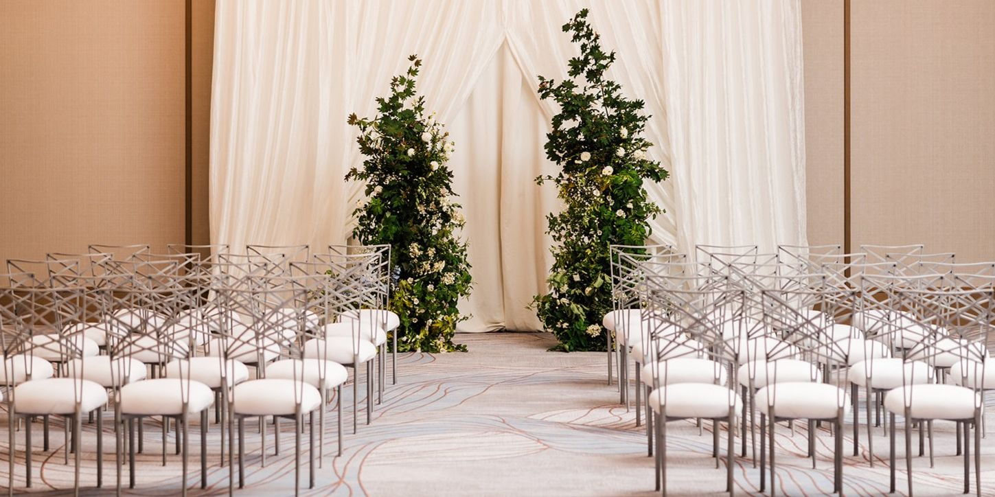 Wedding ceremony setup with rows of white chairs facing a draped backdrop framed by lush greenery and white floral arrangements in an elegant ballroom.