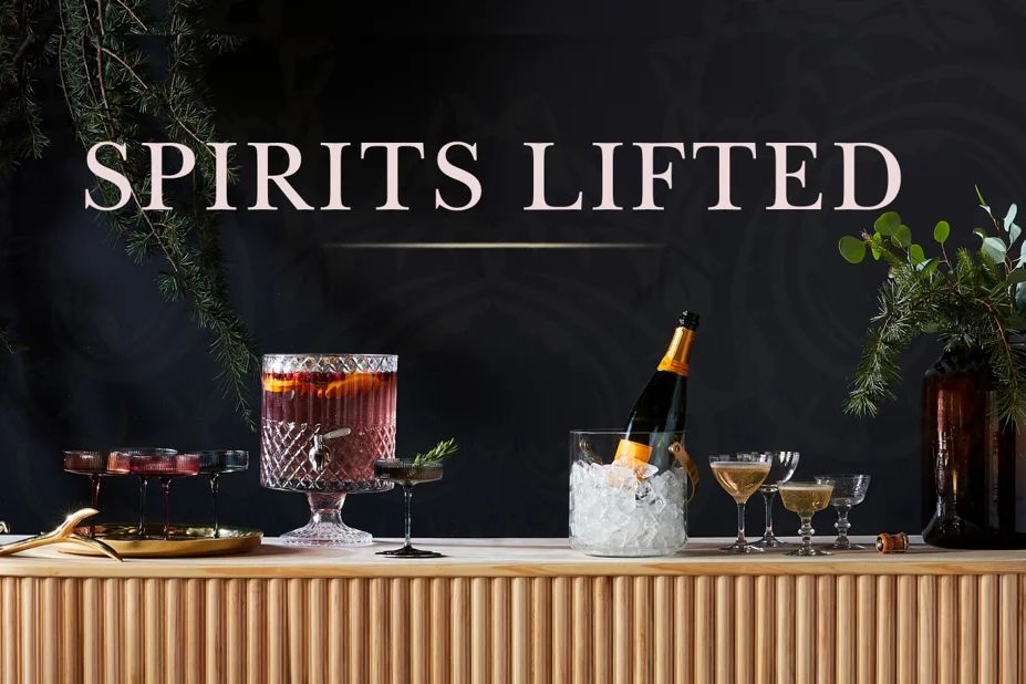 Holiday cocktail display featuring a crystal punch bowl, champagne on ice, assorted stemmed drinks, and seasonal greenery arranged on a bar beneath the text “Spirits Lifted.”