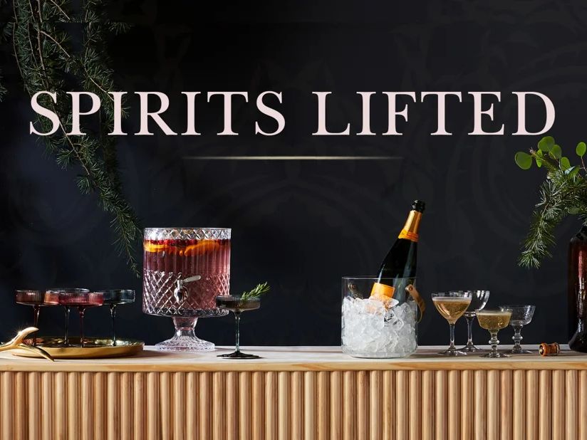 Holiday cocktail display featuring a crystal punch bowl, champagne on ice, assorted stemmed drinks, and seasonal greenery arranged on a bar beneath the text “Spirits Lifted.”