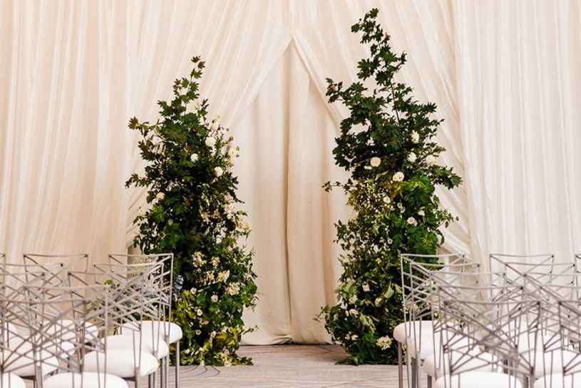 Indoor wedding ceremony aisle with white chairs, draped backdrop, and lush greenery framing the altar space.