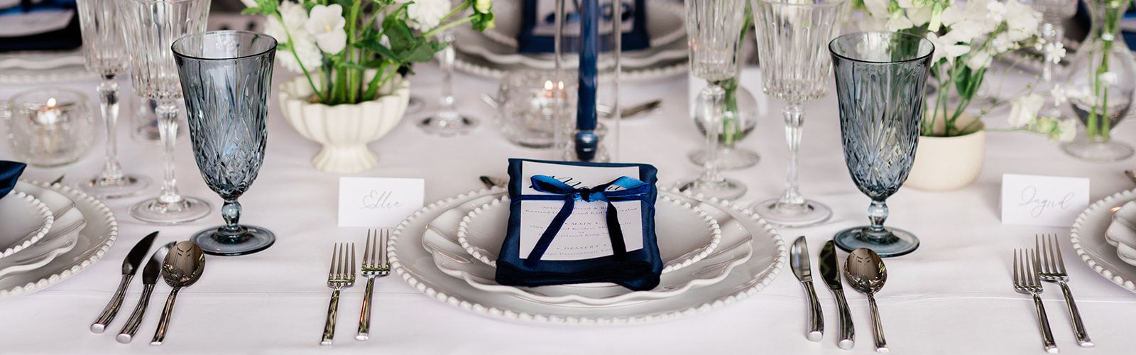 Elegant wedding reception table with white floral centerpieces, blue candle accents, crystal glassware, and place settings with navy napkins and printed menus.