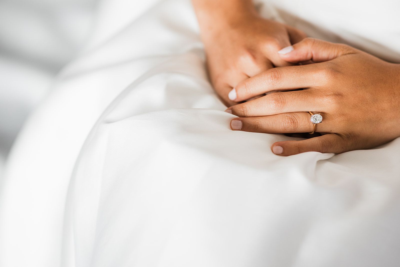 Close-up of hands resting on white wedding attire, featuring a solitaire diamond engagement ring on a gold band.
