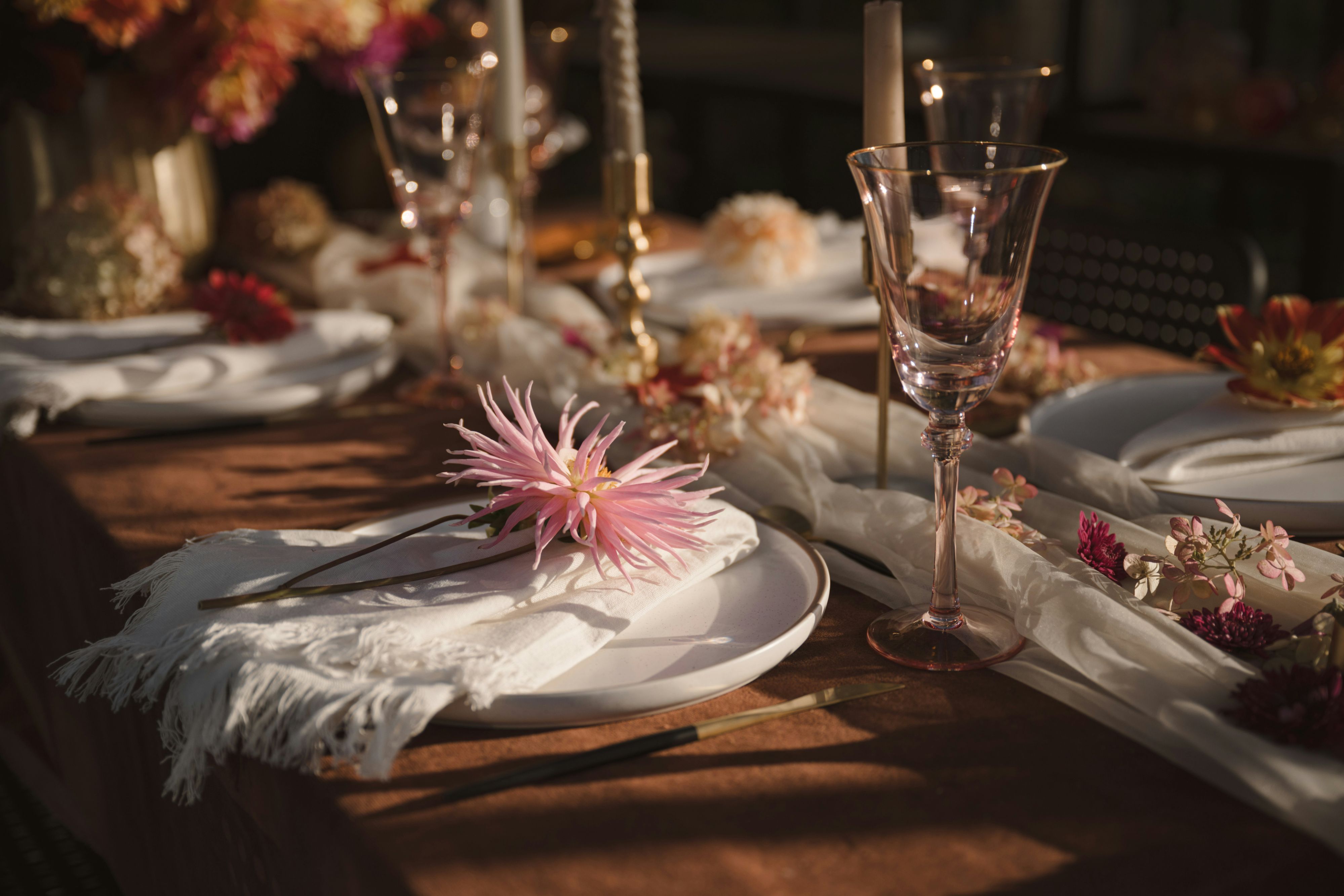Elegant event table setting with white plates, sheer linens, glassware, candleholders, and pink floral accents arranged across a warmly lit dining table.