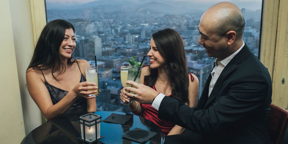 Three guests clink cocktails while seated at a table by a window overlooking the city at dusk.